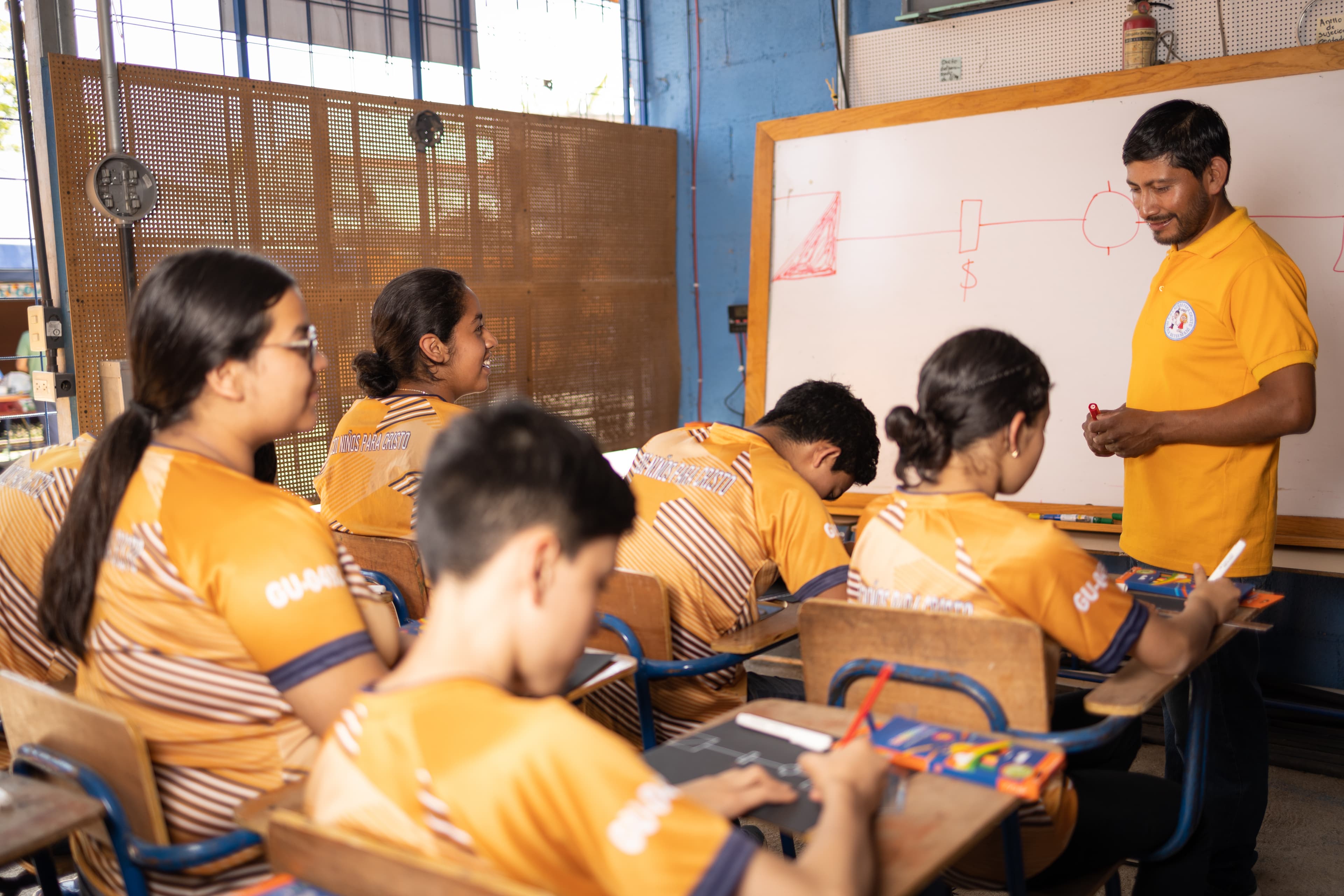 A male tutor stands in front of a whiteboard teaching a group of students at a Compassion center.