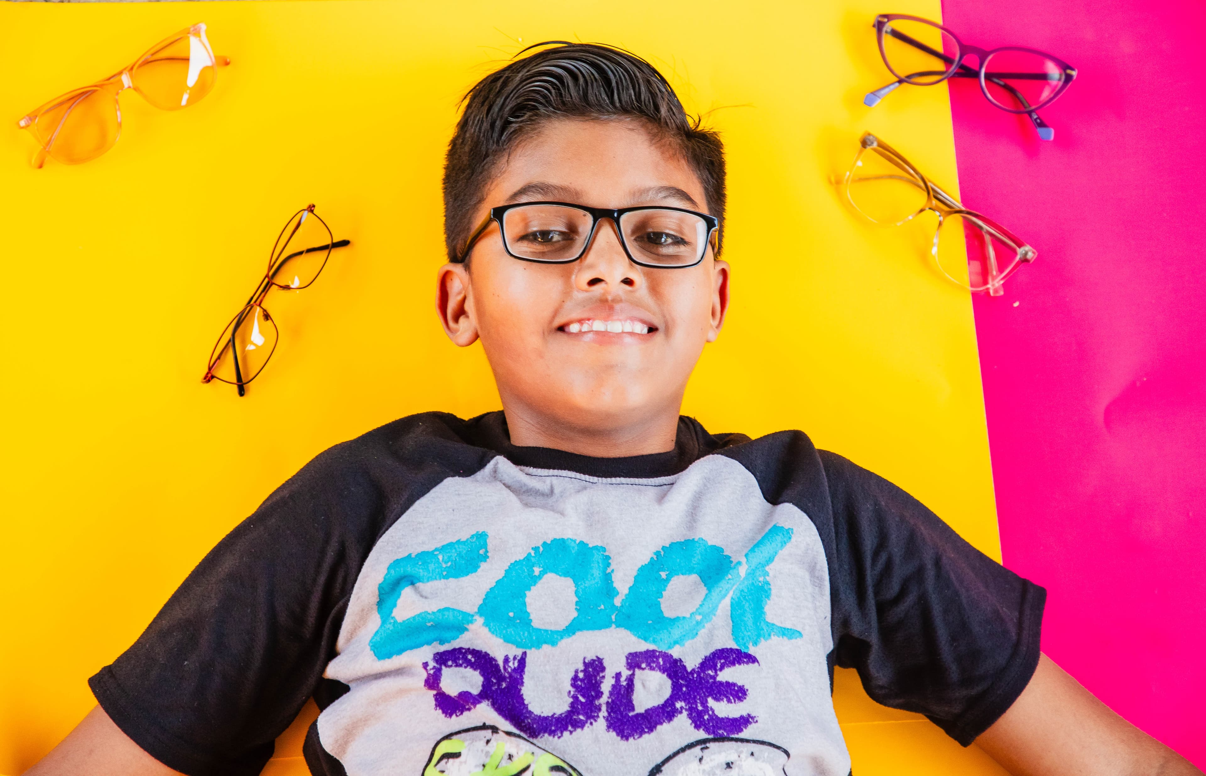 A young boy wearing glasses lies on a yellow and pink background.