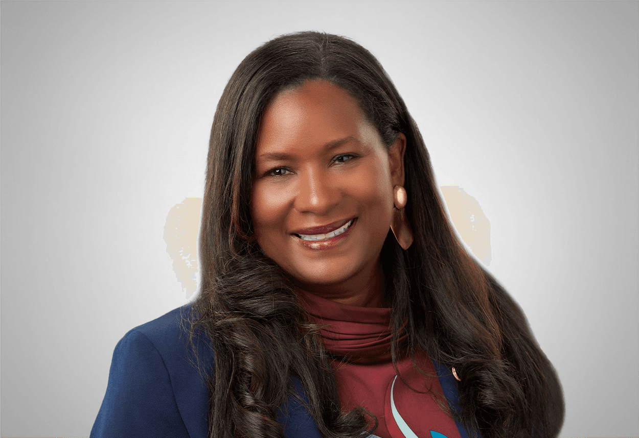 A woman in a maroon printed blouse and blue blazer smiles in front of a black background.