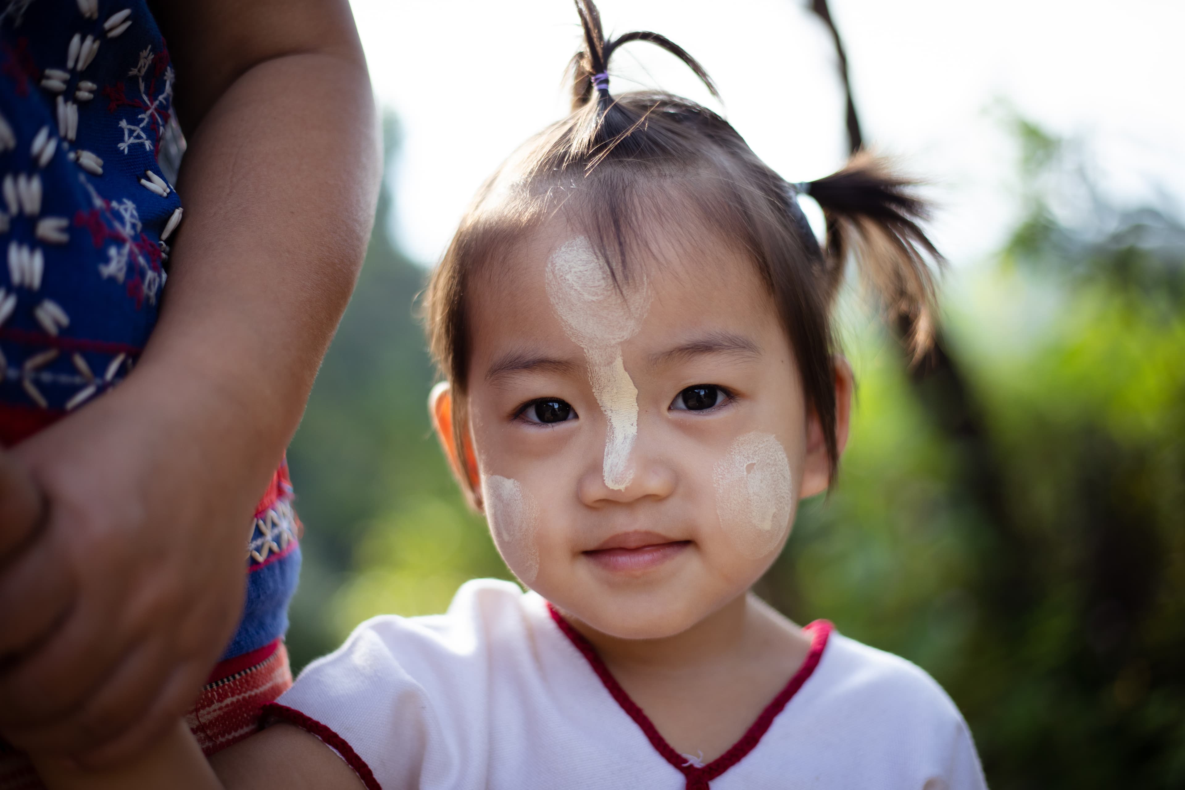 A close-up shot of a young girl’s smiling face. She has paste on her face. She is holding an adult’s hand.