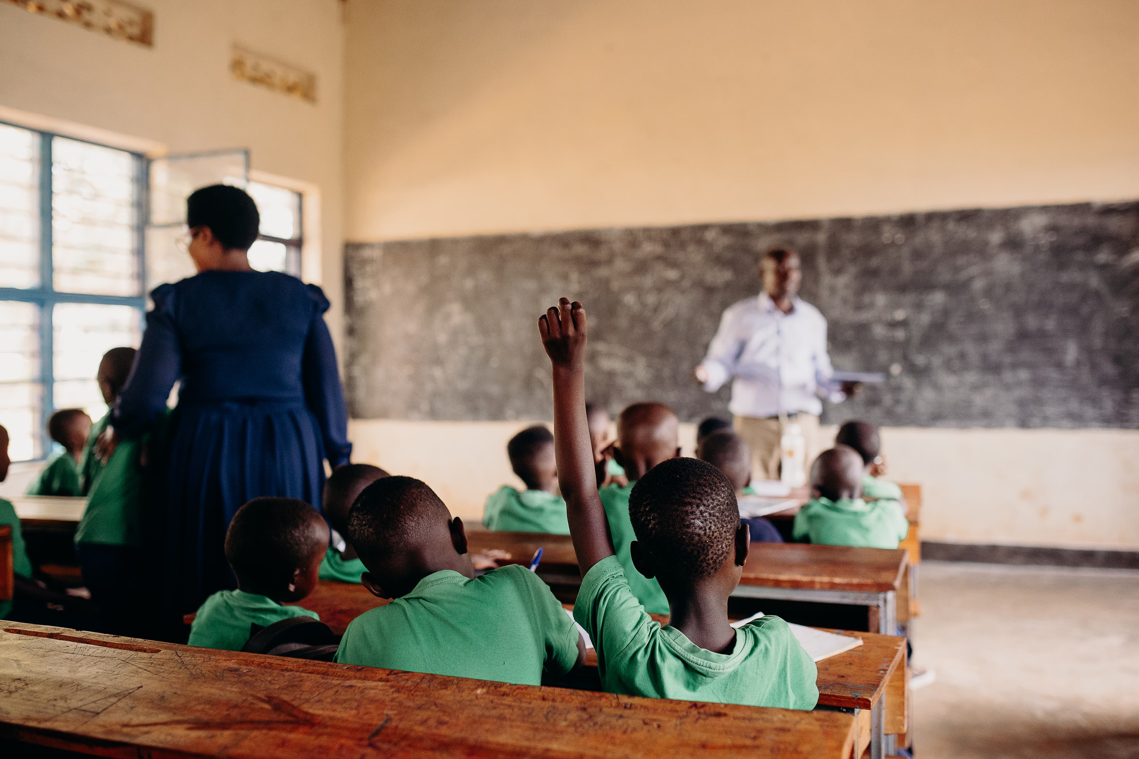 Young boy is sitting at desk raising his hand as teacher stands at the chalkboard in front of him.
