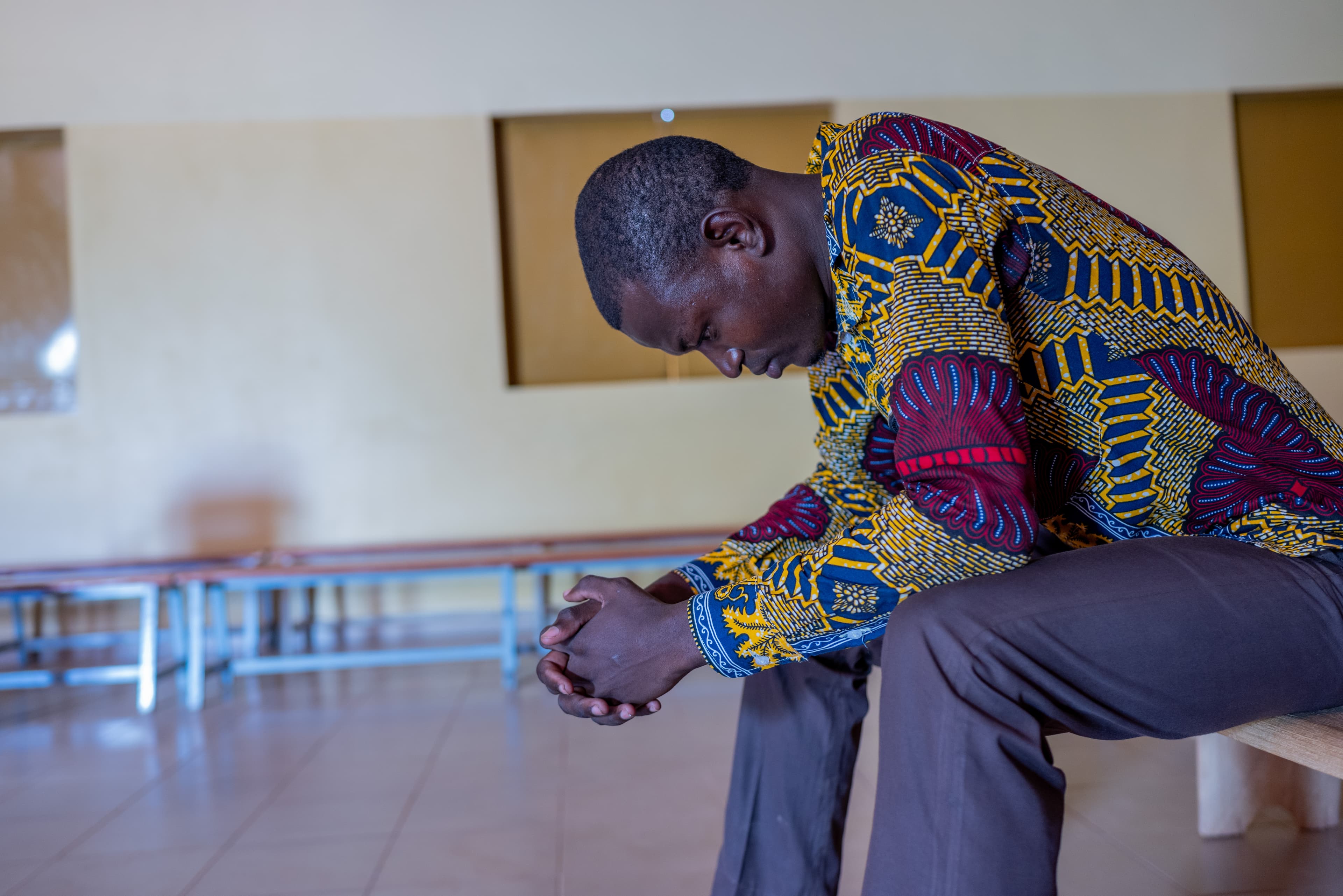 An African man sits on a wooden chair and kneels forward in prayer.