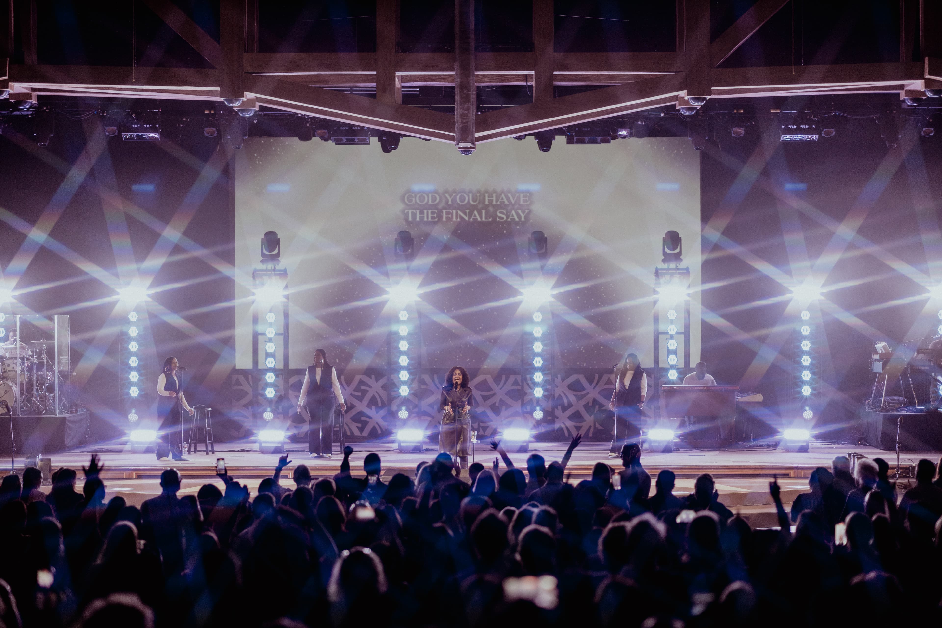 A woman stands on a stage with backup singers, with bright lights and text on a screen in the background.