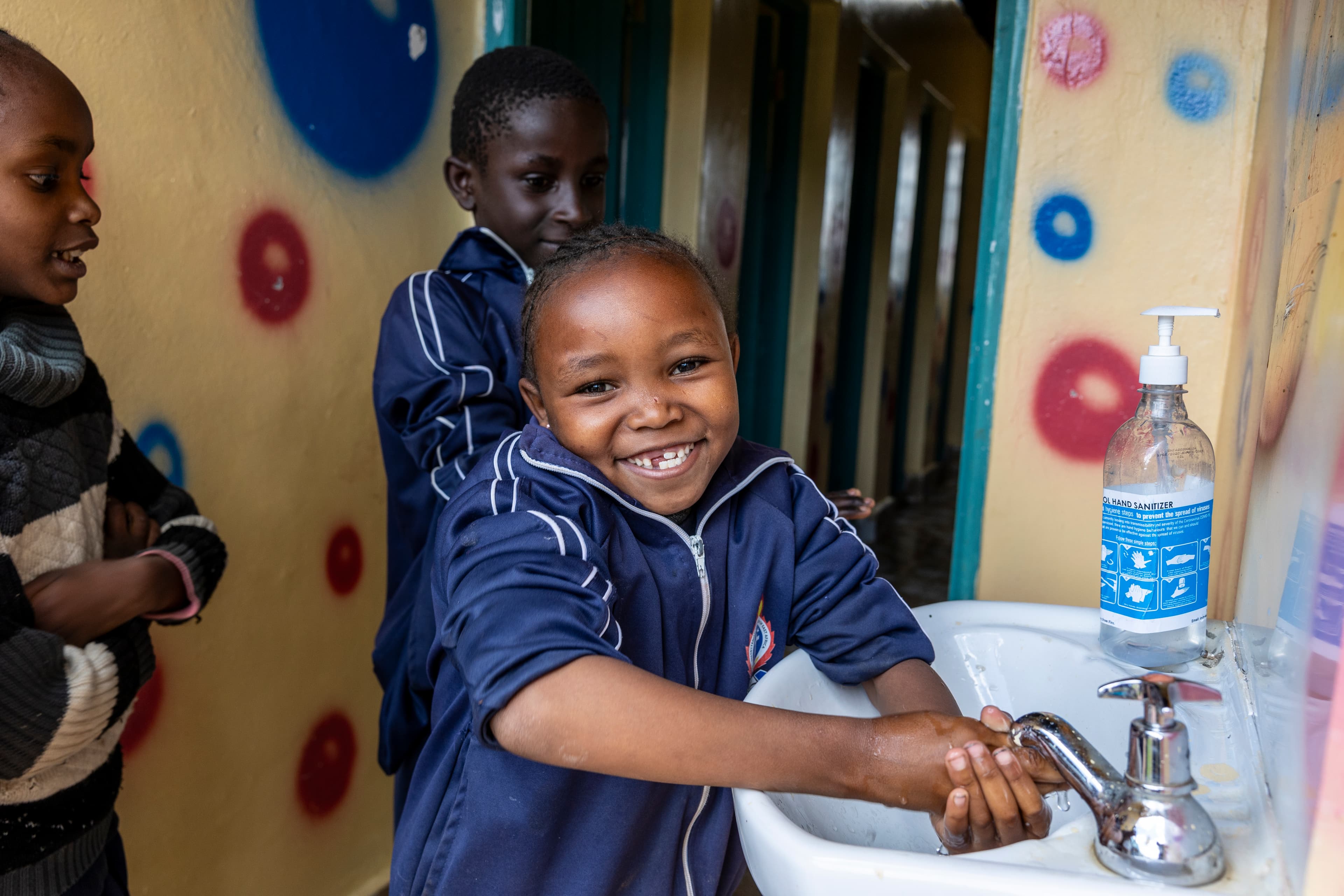 A young girl washes her hands in a white sink as she smiles for the camera.