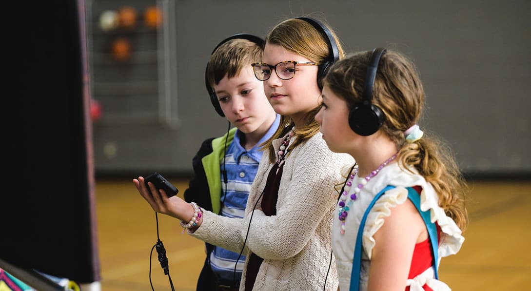 To girls and a boy wear black headphones, listening to a black electronic device that the girl in the middle is holding. They are standing in a gymnasium.