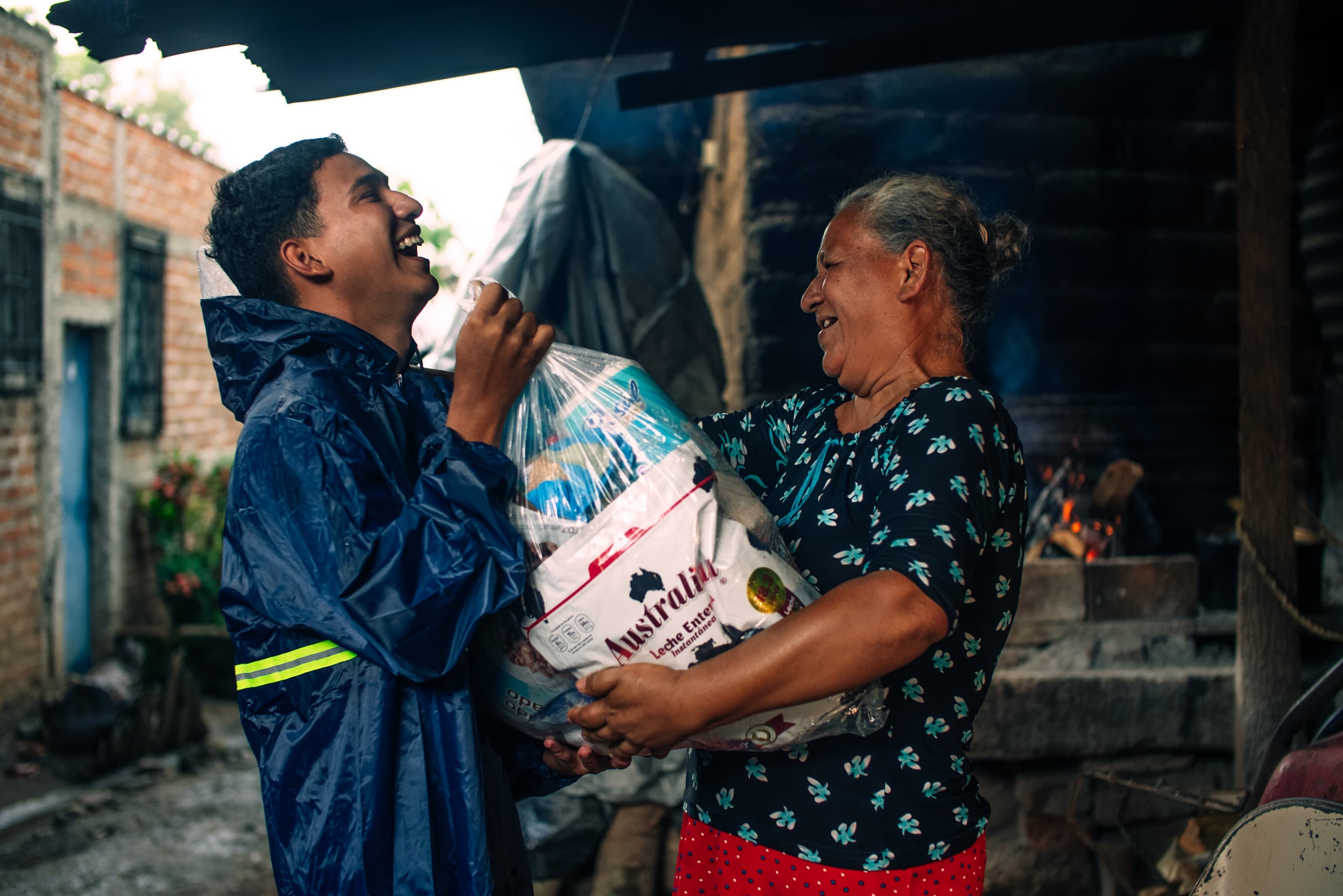 A young man wearing a raincoat gives a bag of food and supplies to an older woman. They are both smiling.