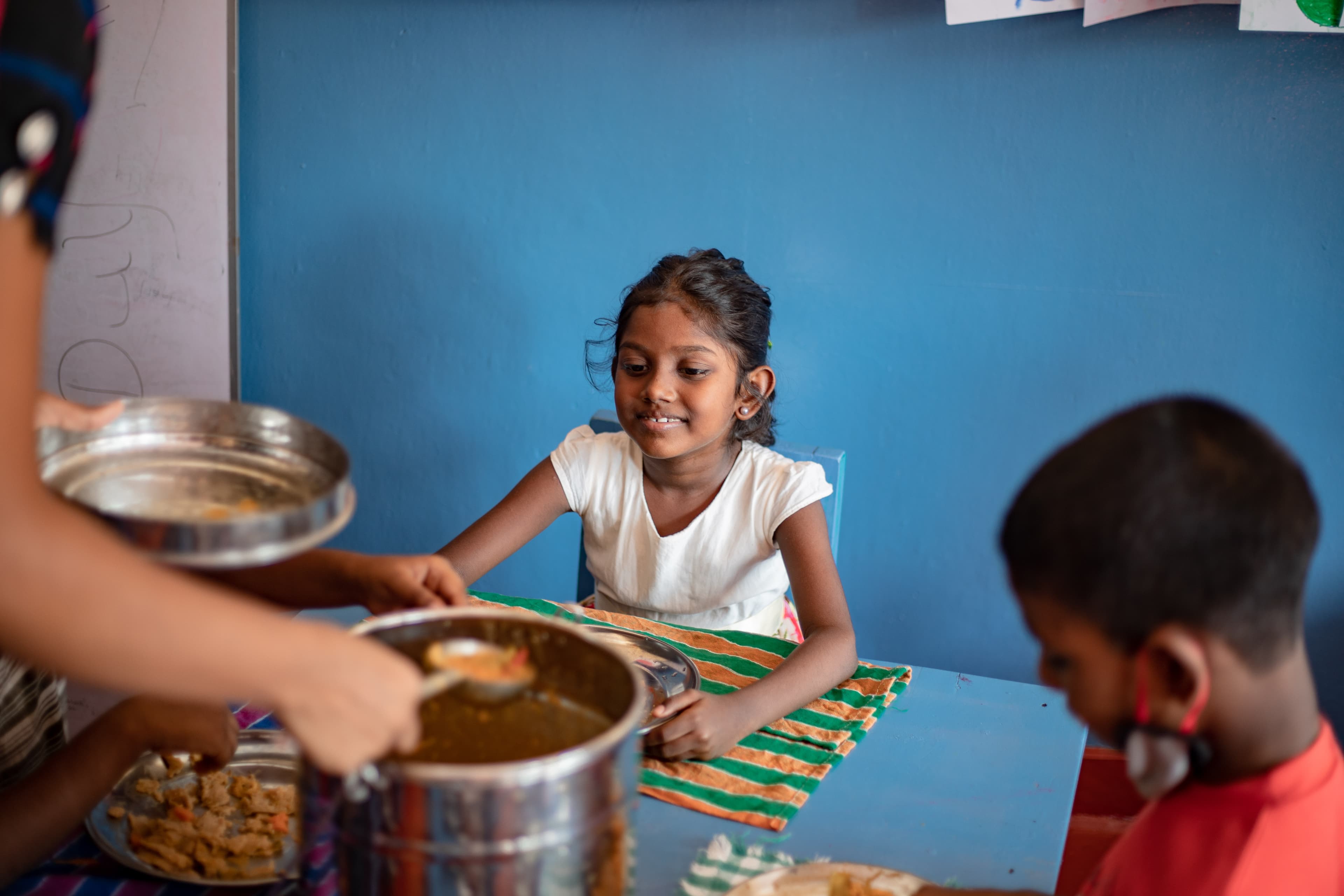 A young girl in a white shirt awaits her food, sitting at a blue table.