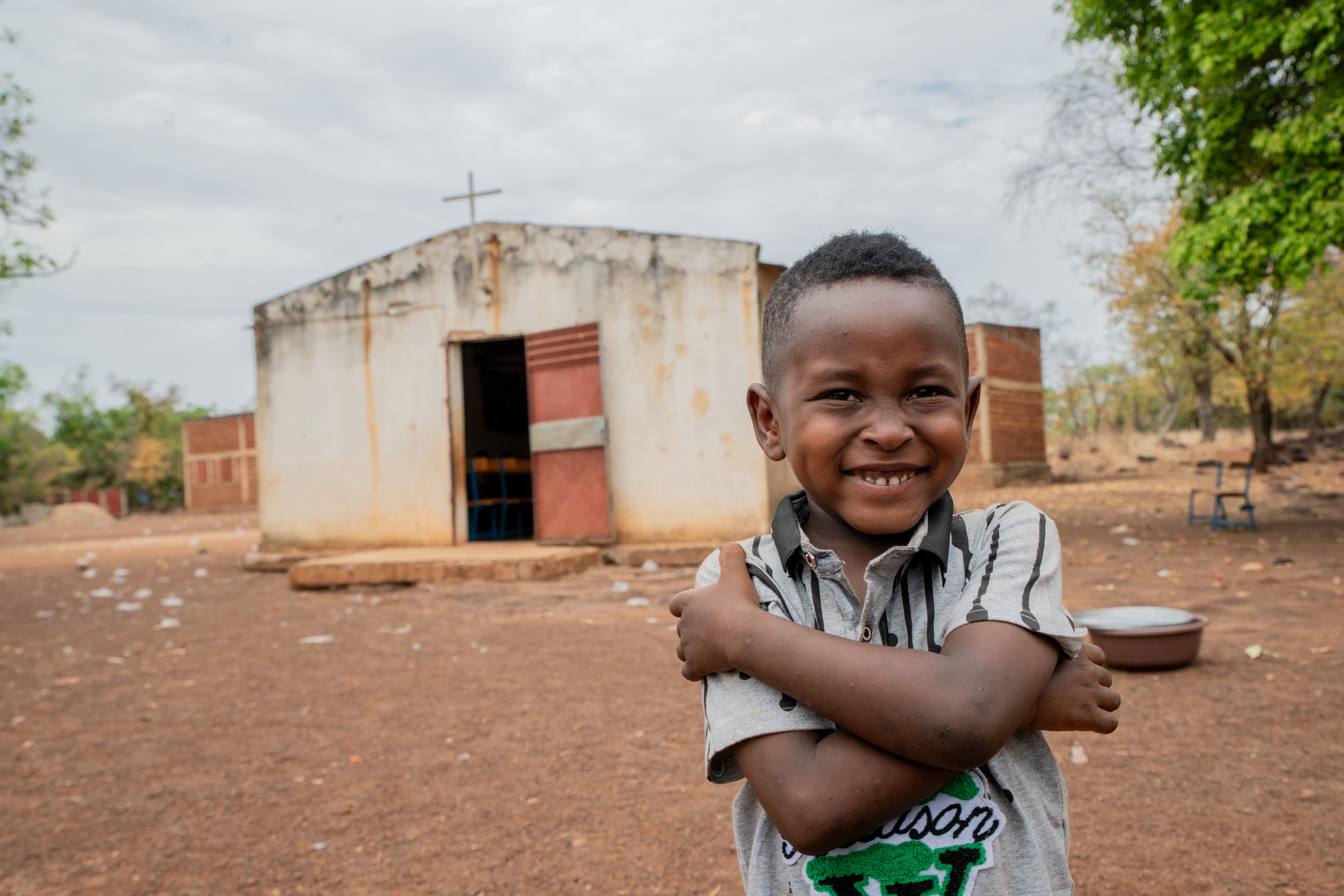 A young African boy crosses his arms and smiles for the camera.