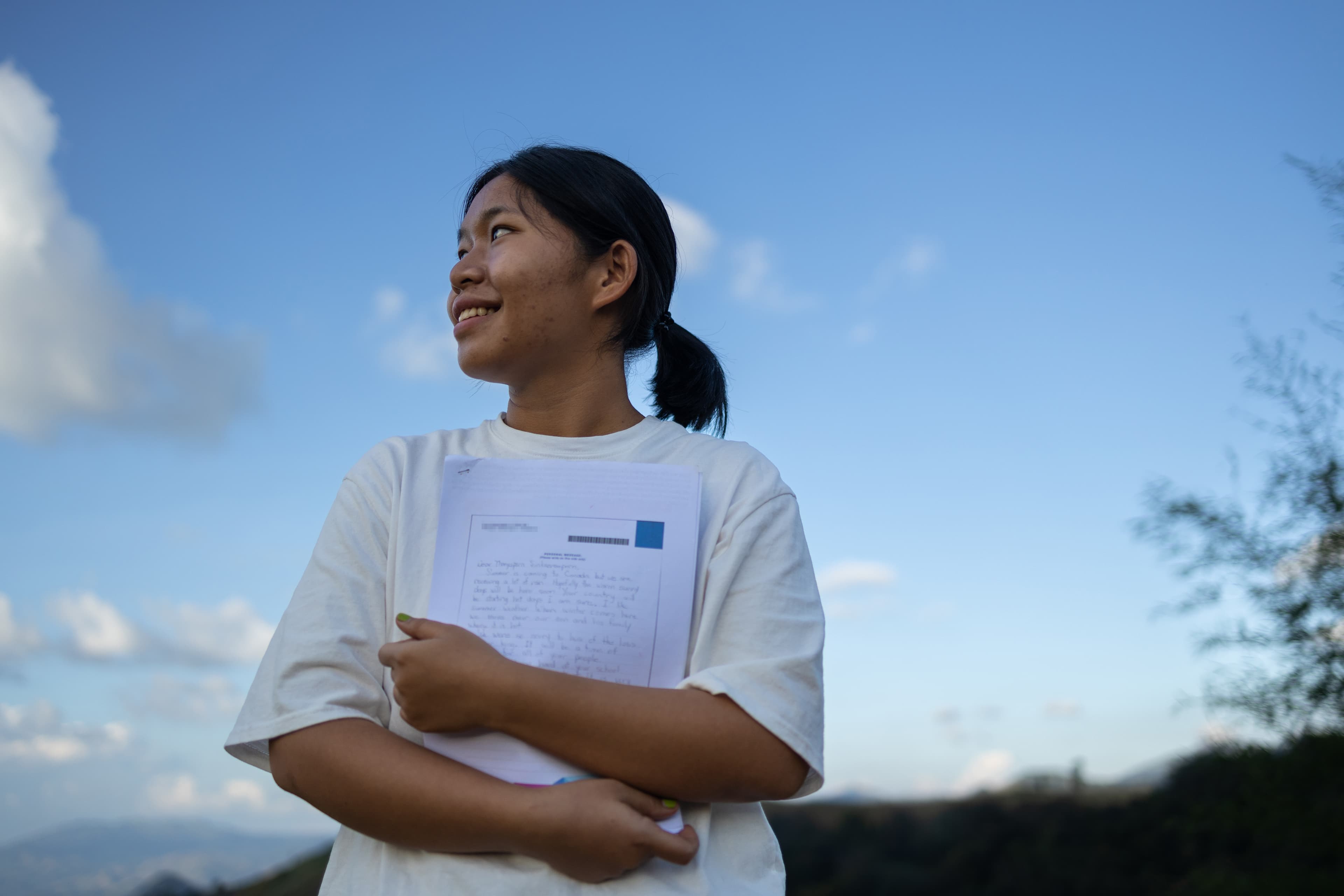 A young woman is standing and holding her sponsor letter looking into the distance.