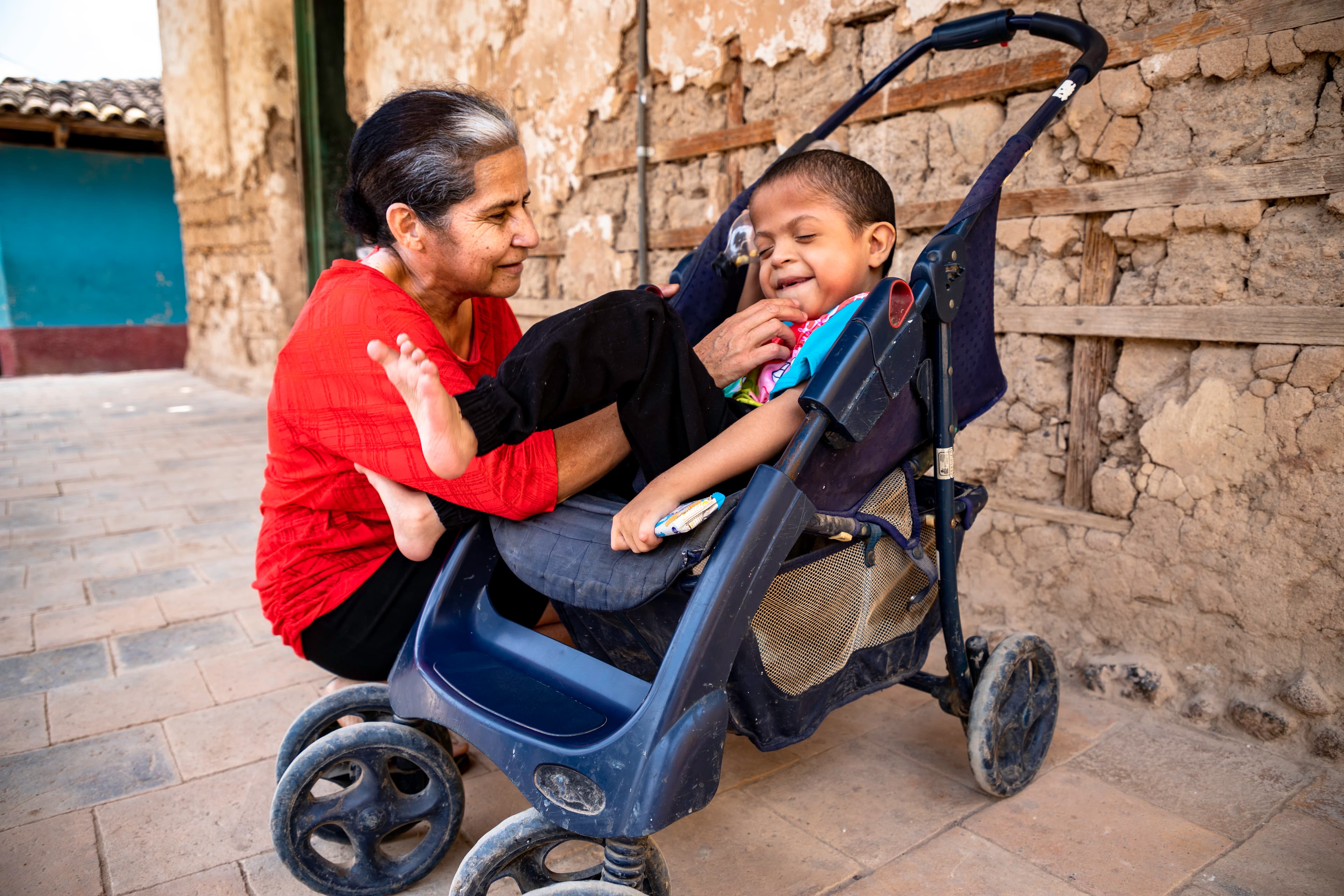 A little boy is sitting in his stroller outside his home next to his mother who is tickling him.