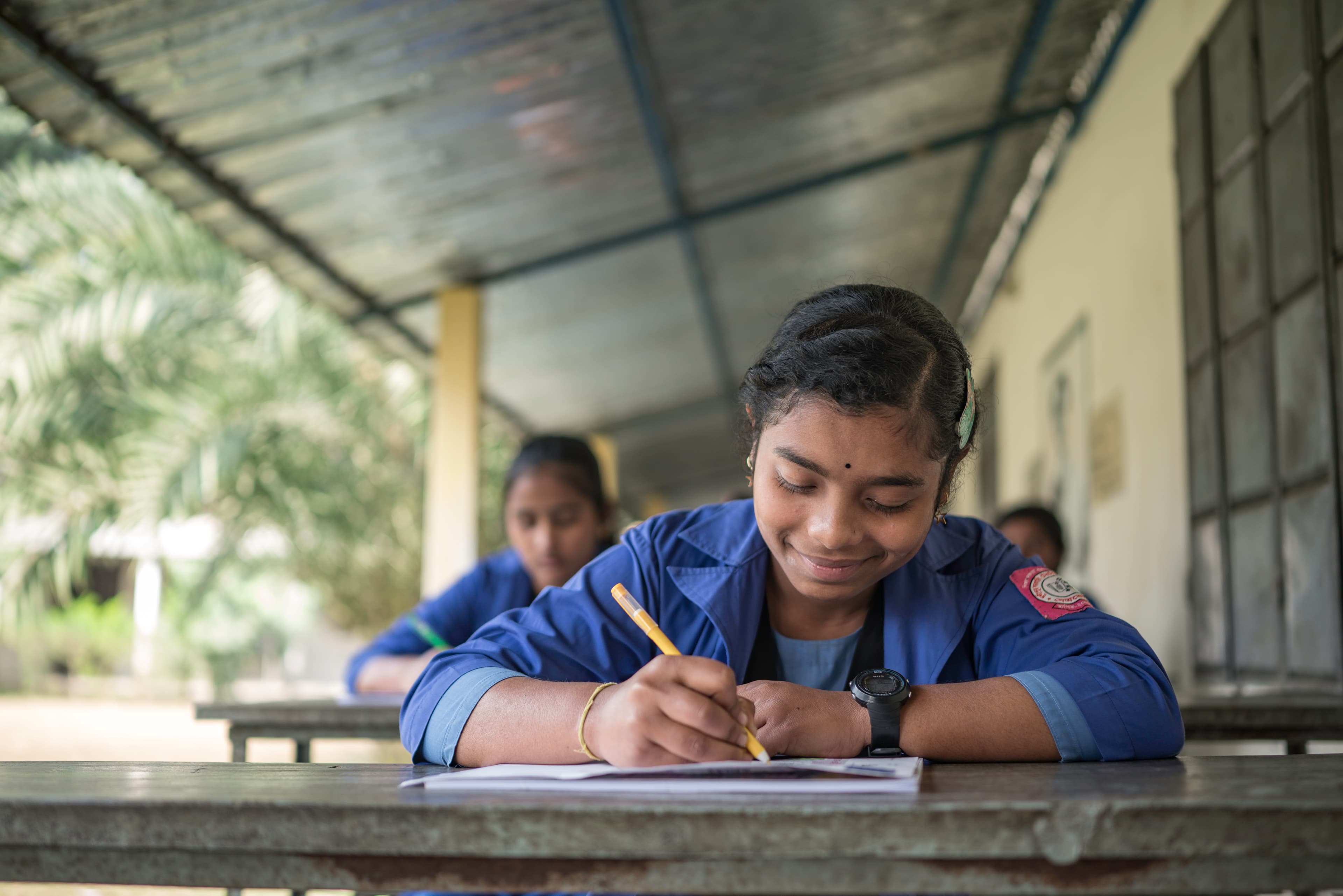 A young woman is sitting at her desk outdoors writing a letter with a smile on her face.