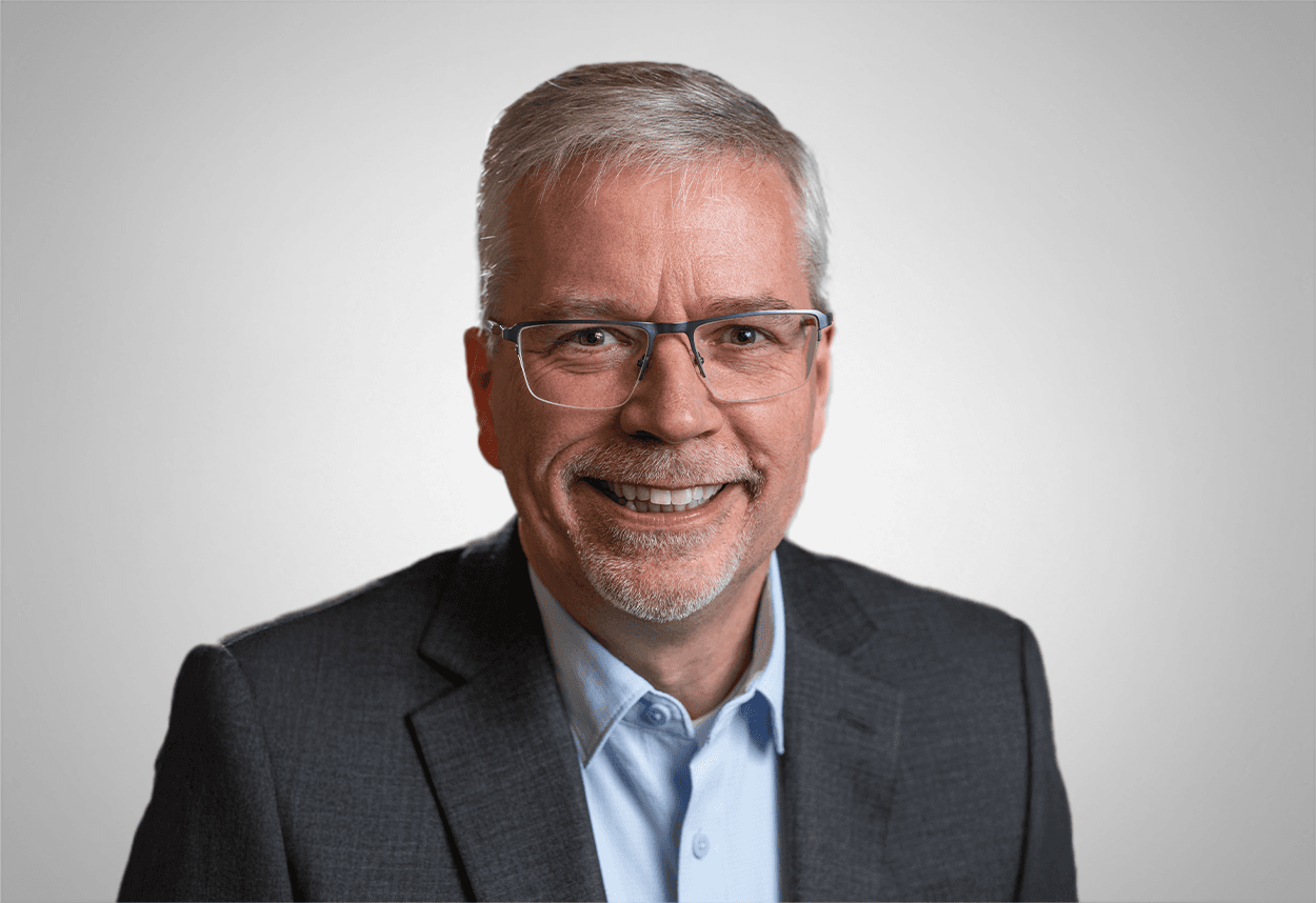 A man wearing a dark grey suit jacket and glasses is smiling, with a grey background behind him.
