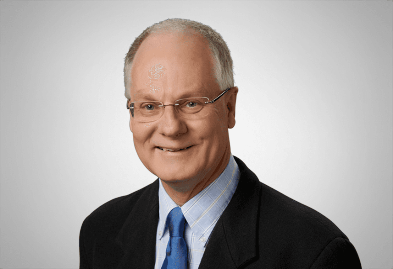 A man wearing a light blue checkered shirt, blue tie and black suit jacket is smiling, with a grey background behind him.
