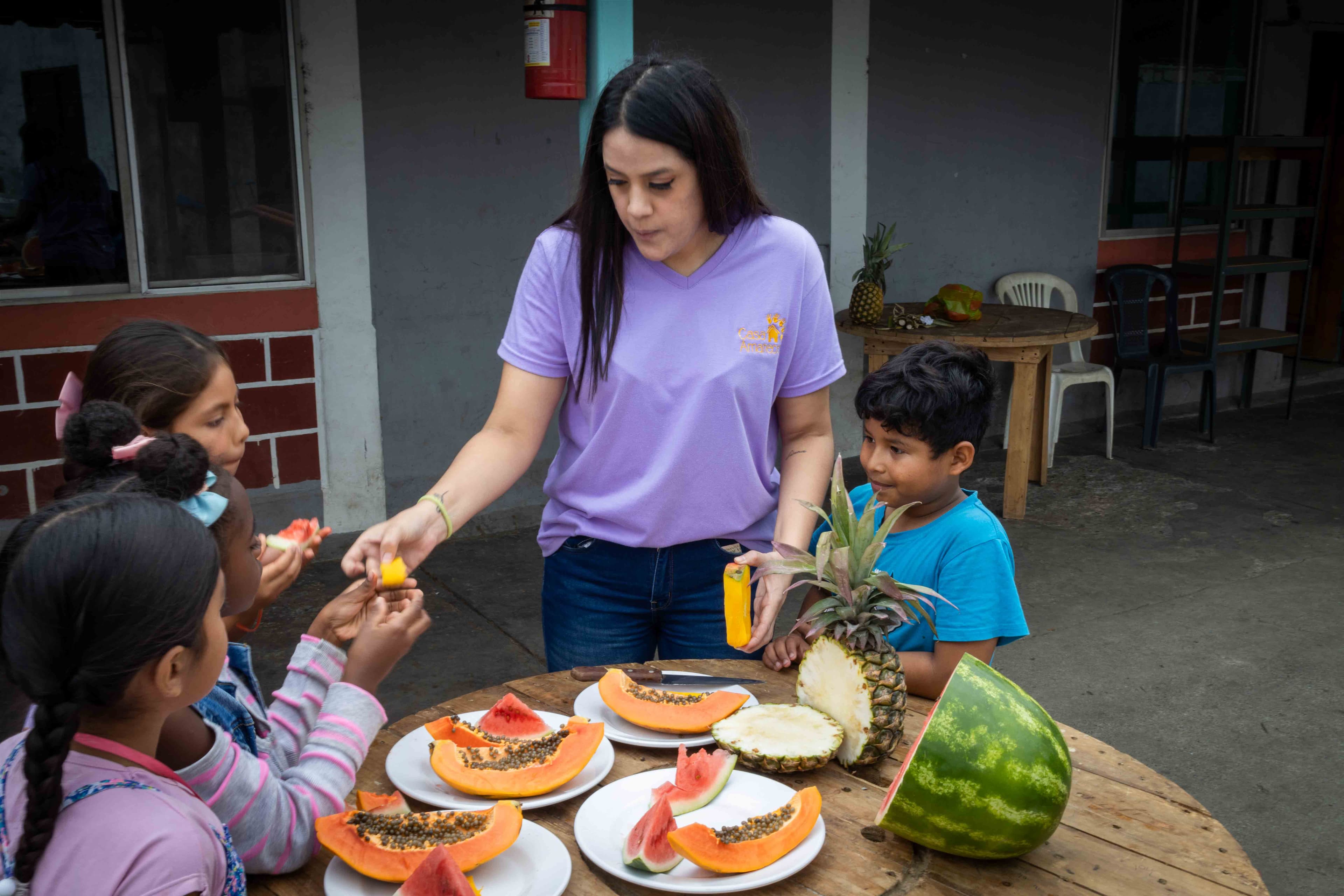 An adult woman shares fruit with a group of children.