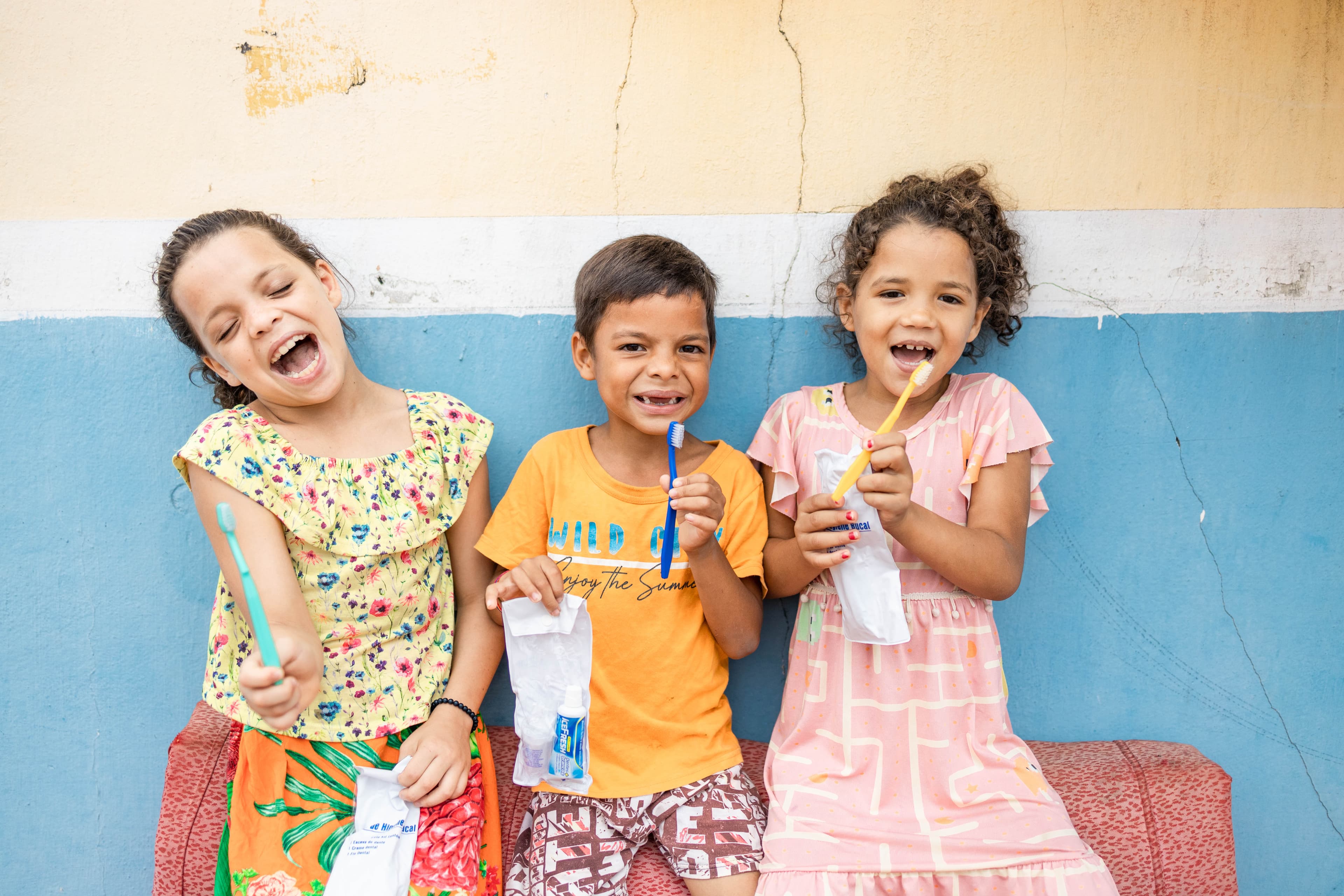 Two young girls and a young boy in colorful clothing smile and laugh while holding