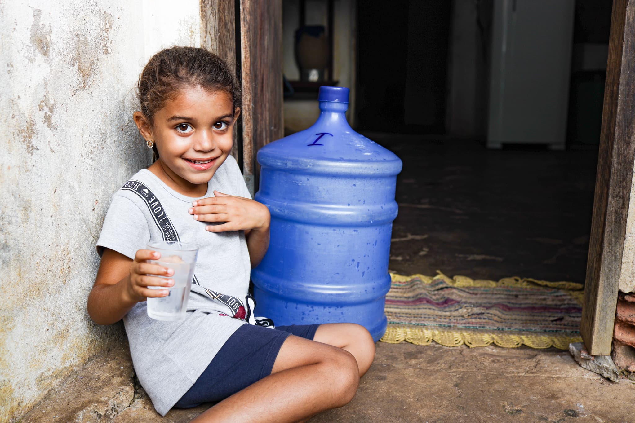 Young girl smile as she sits on the ground next to a large bottle of filtered water, holding a glass of clean drinking water.