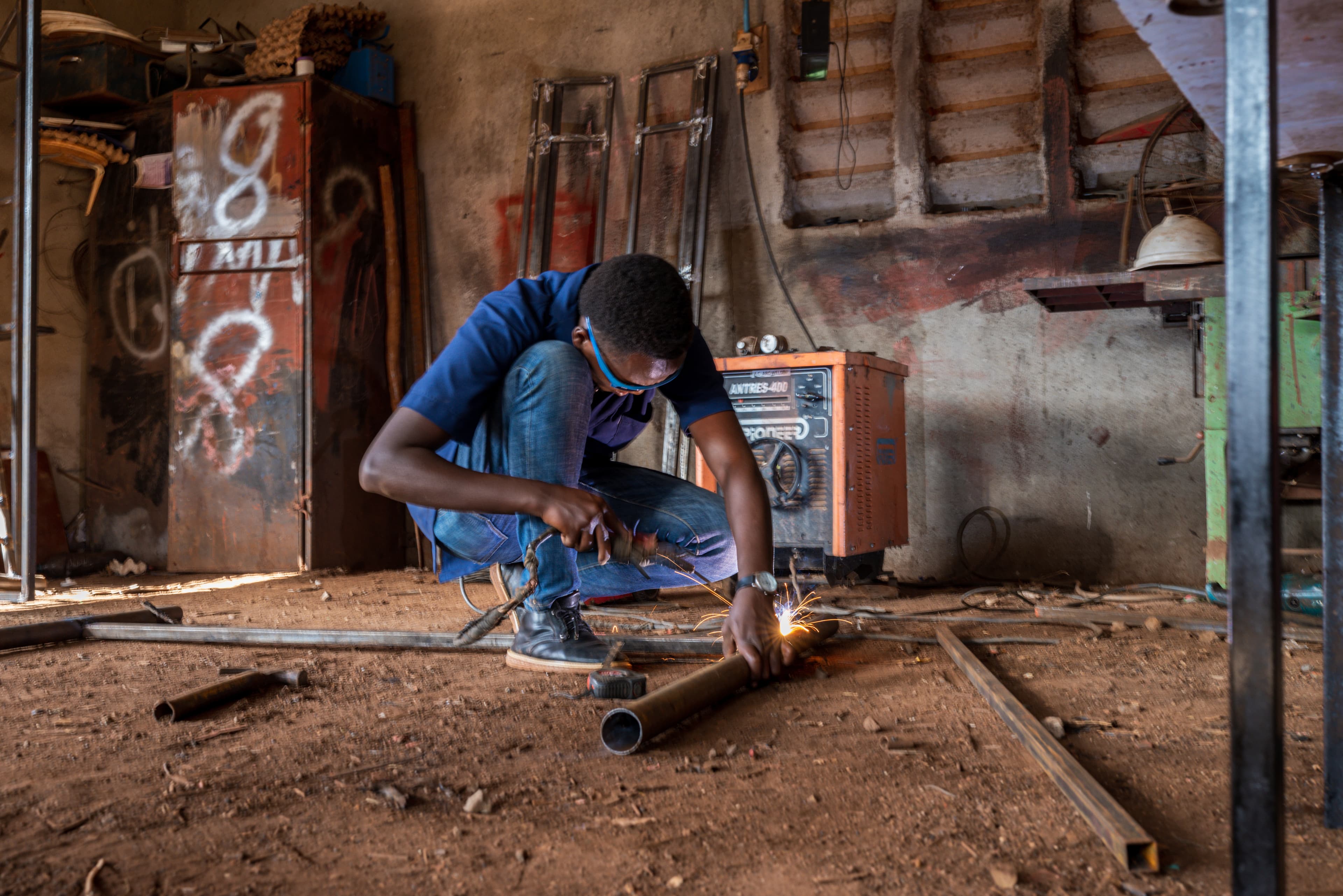 A young boy is leaning down in the workshop as he welds metal.