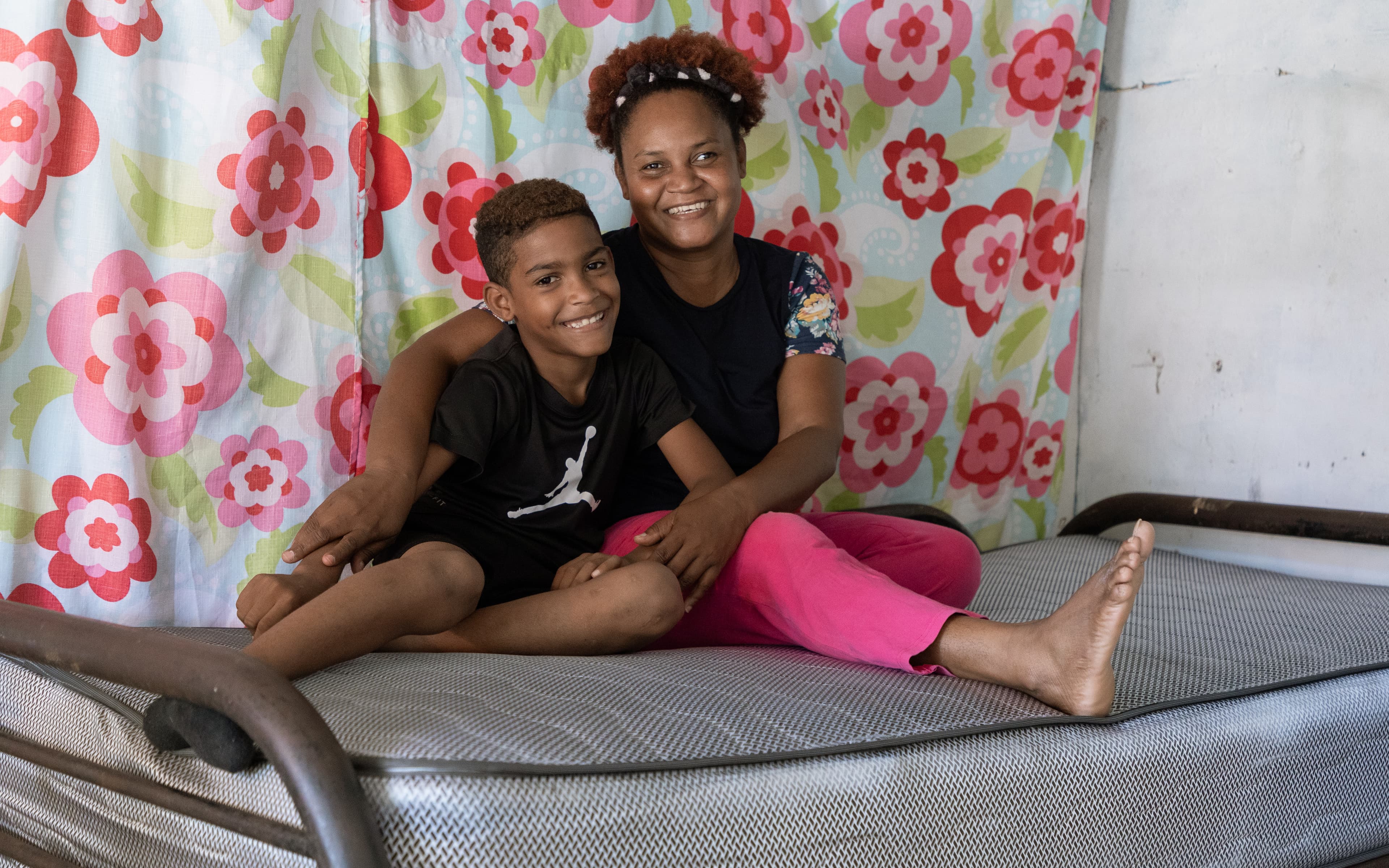 A young boy and his mother sit together on the top mattress of a bunk bed, smiling at the camera.