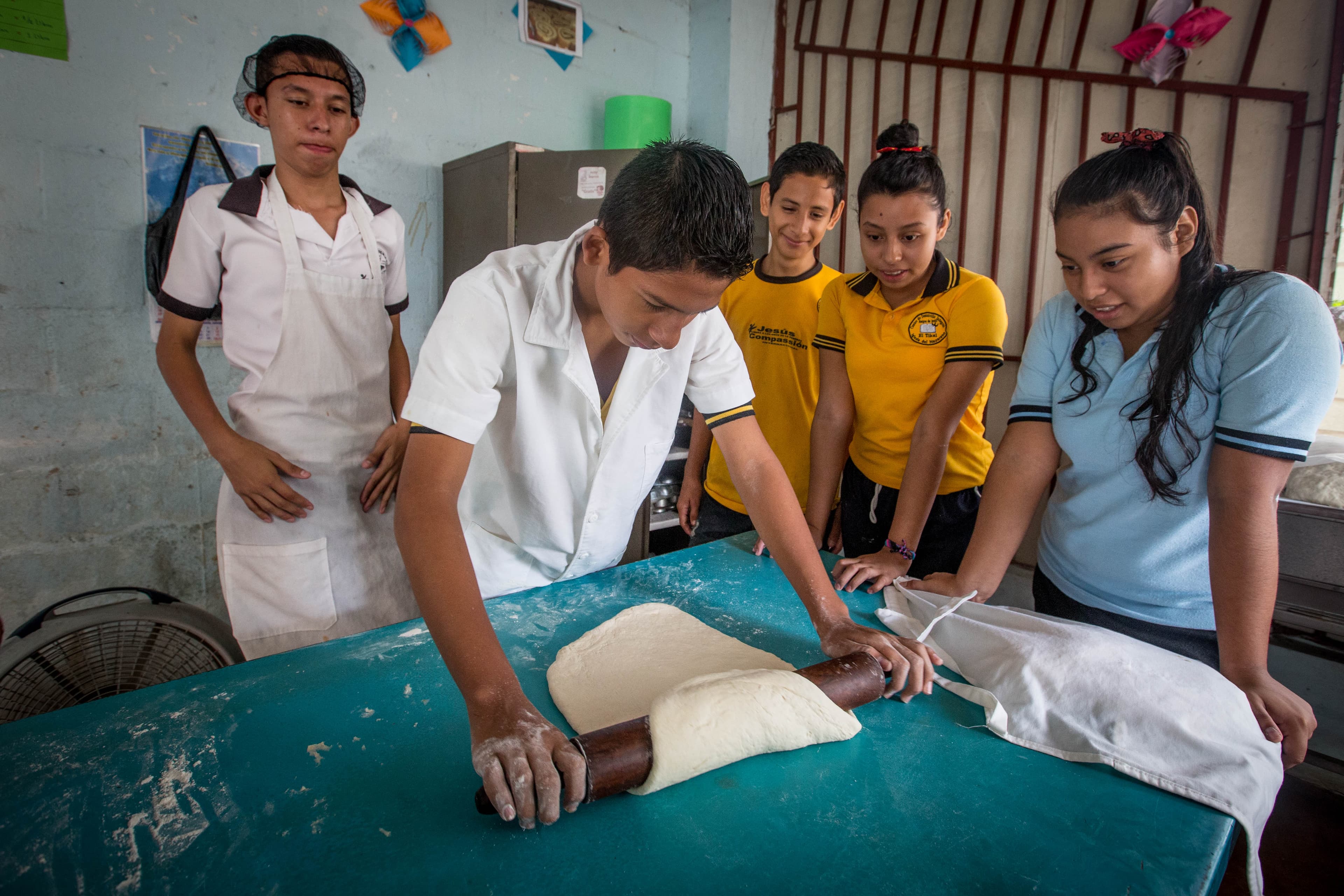 A group of teenagers observe dough rolling during a cooking class.