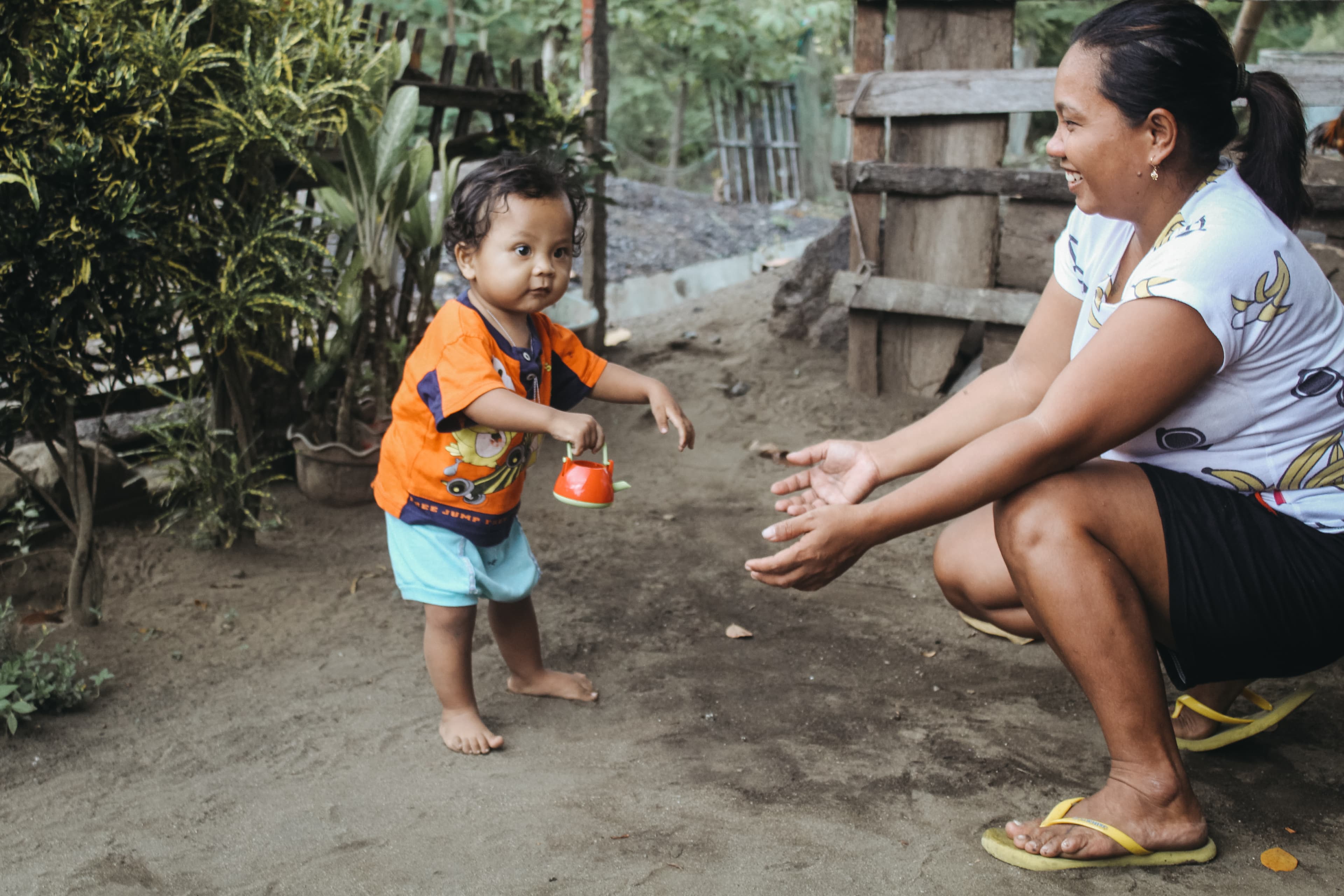 A toddler is wearing light blue shorts and an orange shirt. His mom is wearing black shorts and a white shirt. She is kneeling down with her arms outstretched helping the boy walk. Behind them is a fence.