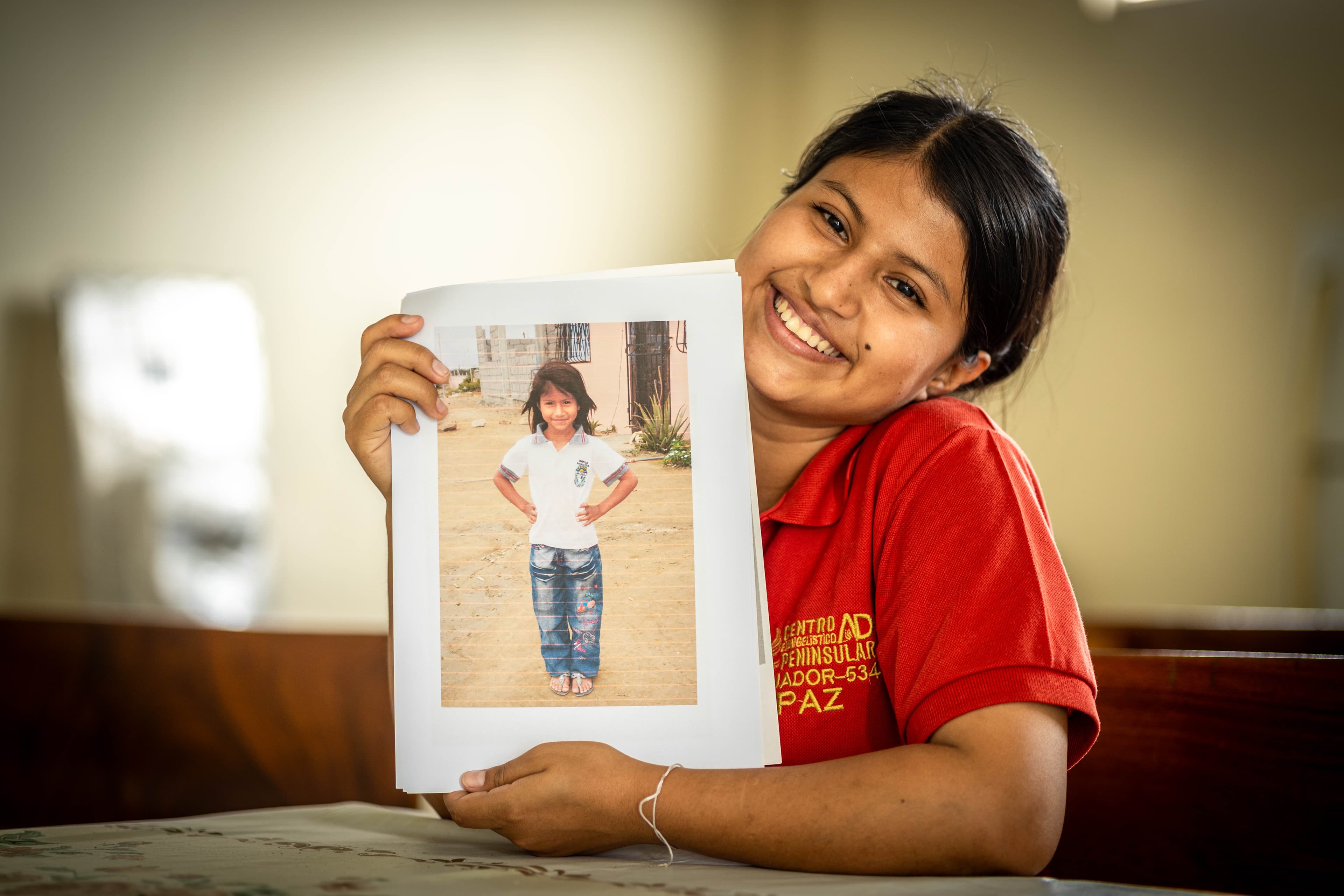 A young woman in a red shirt sits at a table, holding a photo of herself as a child.