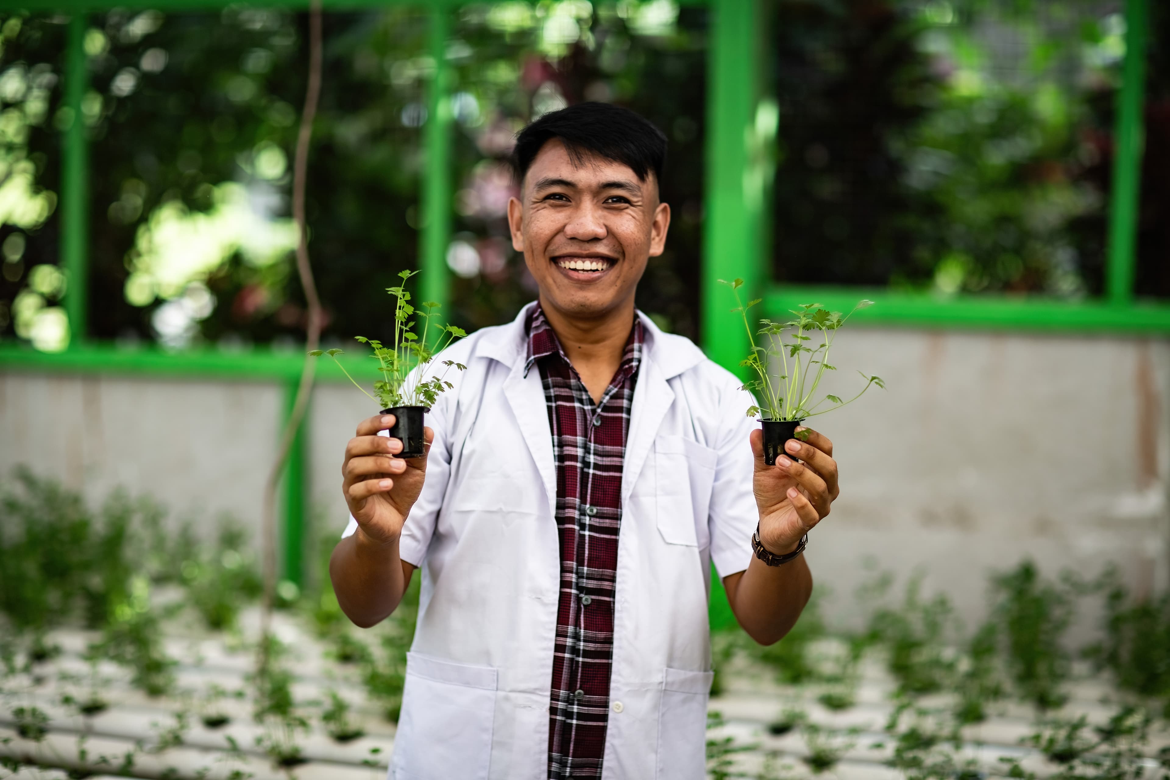 A young man wearing a white lab coat holds plants in his hands while smiling for the camera.