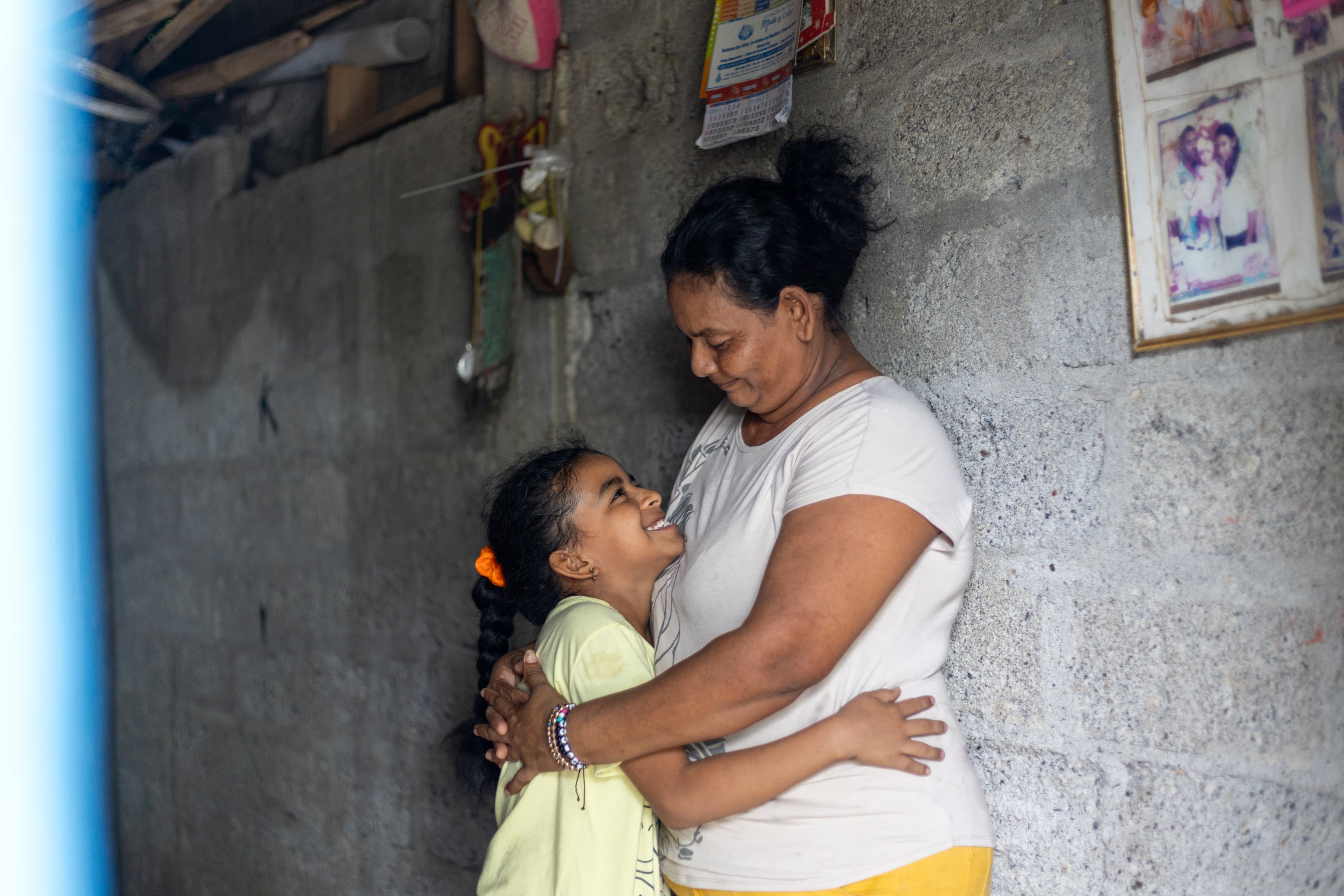 A girl hugs her grandmother, standing in front of a cement wall in their home.