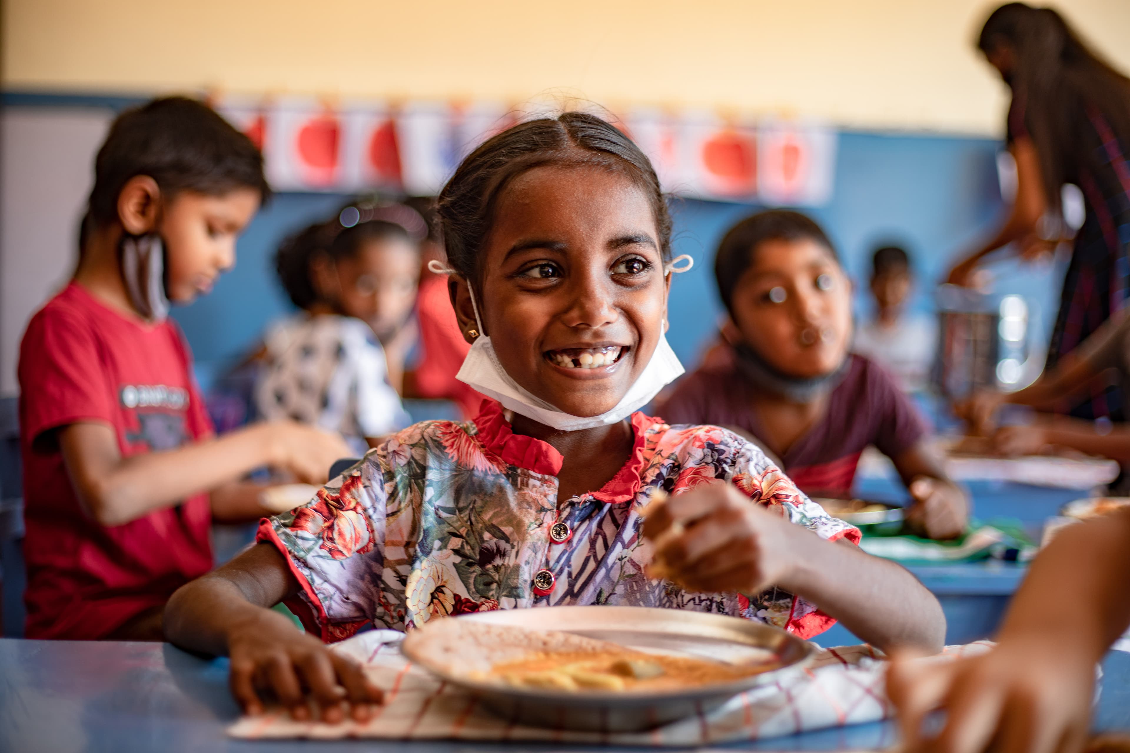 A young girl smiles as she sits down to eat a plate of food surrounded by classmates.