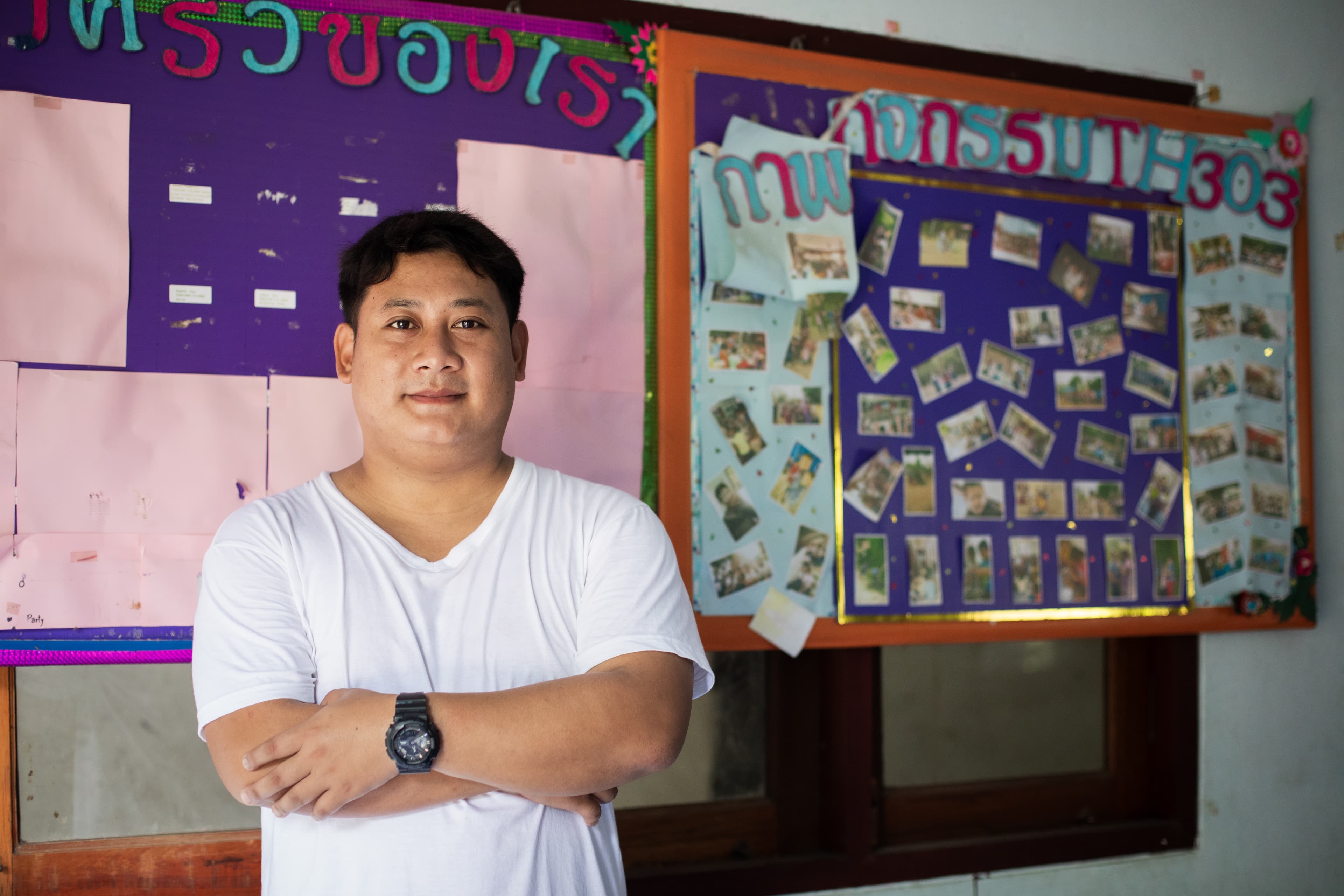 An older man wearing a white t-shirt stands with his arms crossed and smiles for the camera.