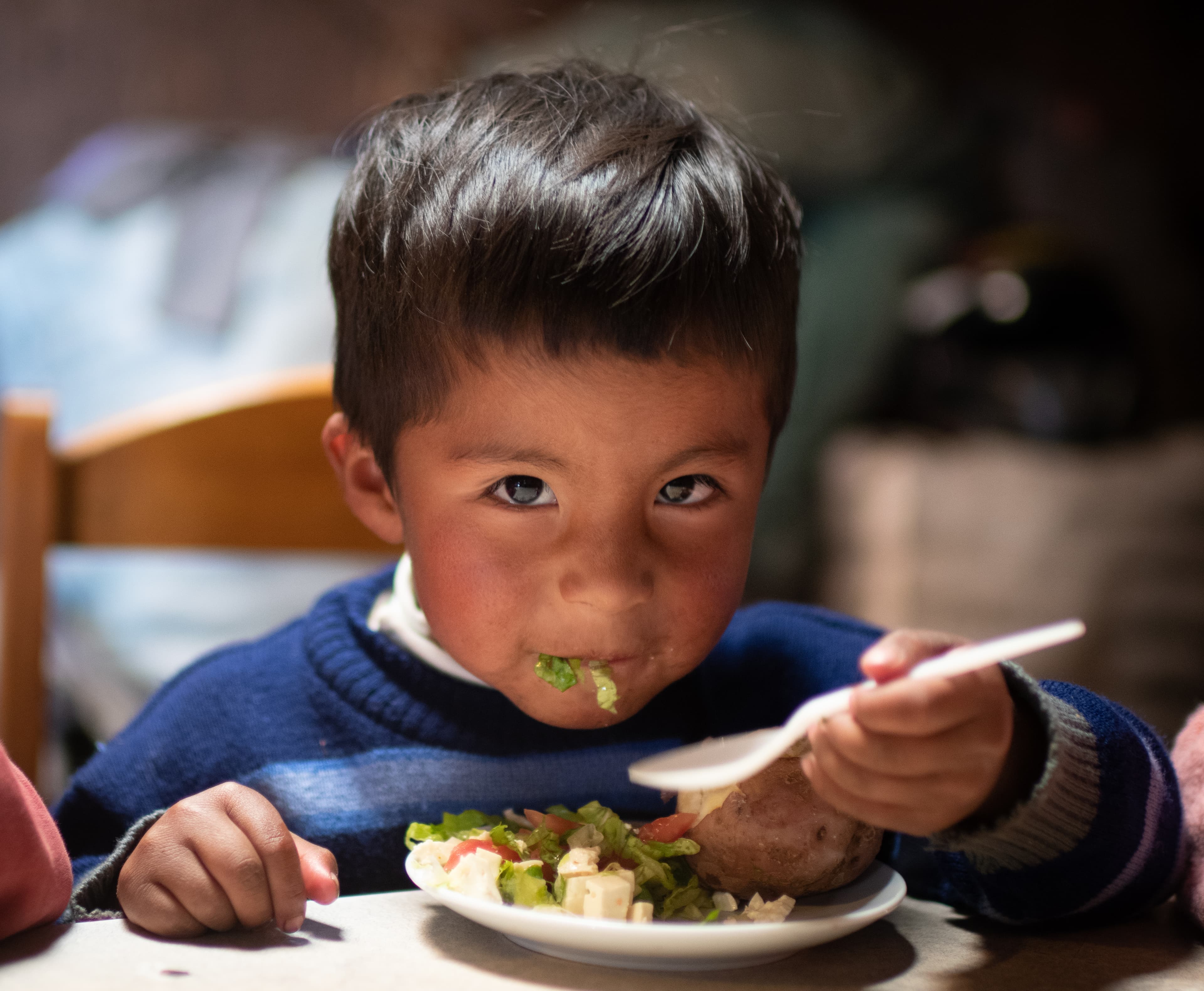 A young boy is looking at the camera while he sits in a chair eating his meal.