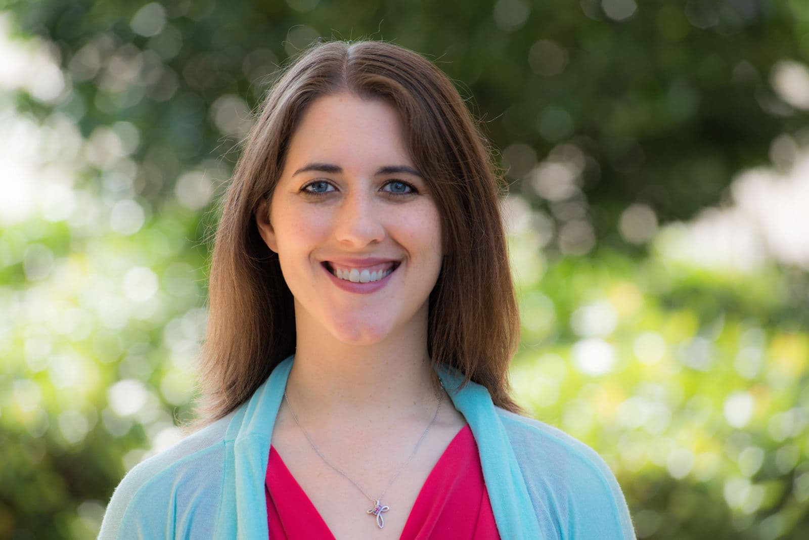 A woman wearing a light blue sweater over a pink top smiles, with trees in the background.