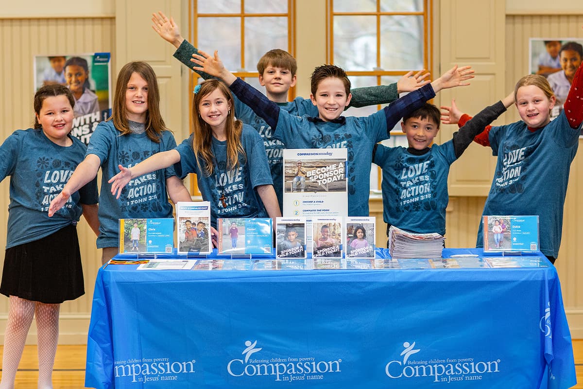A group of young students stand behind a Compassion table smiling with their hands in the air.