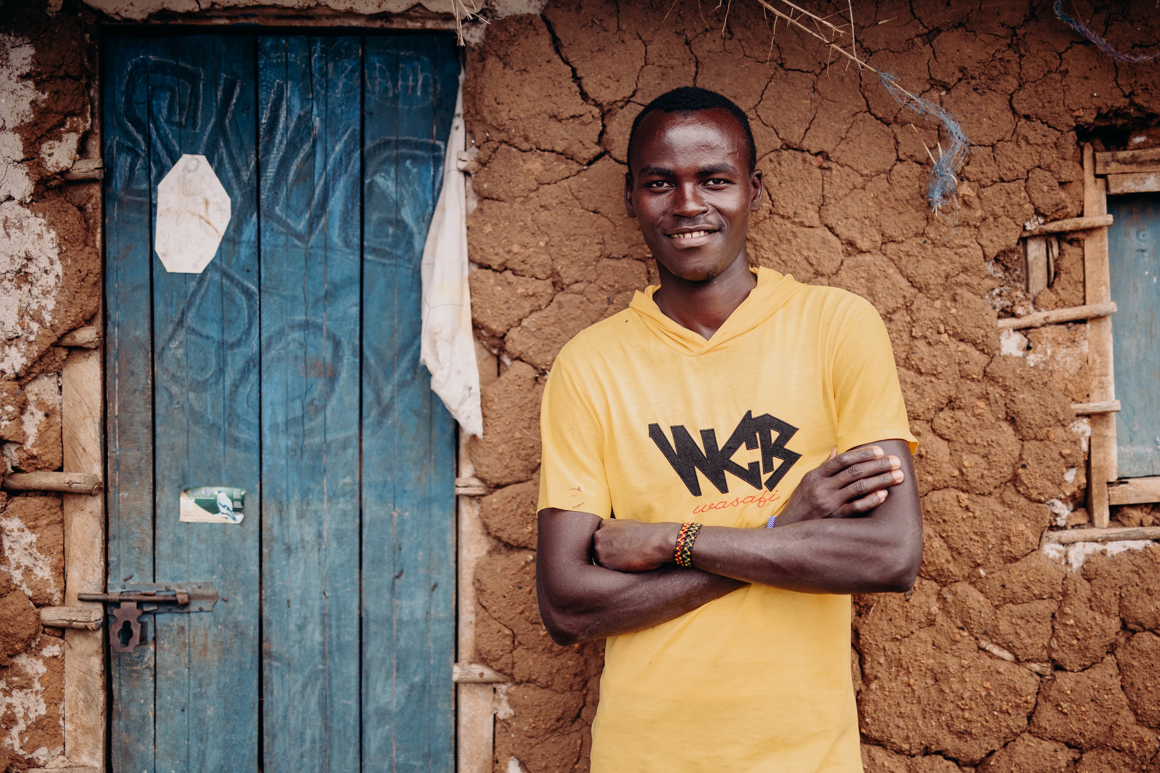 An African teen wearing a bright yellow shirt stands outside of a clay home.