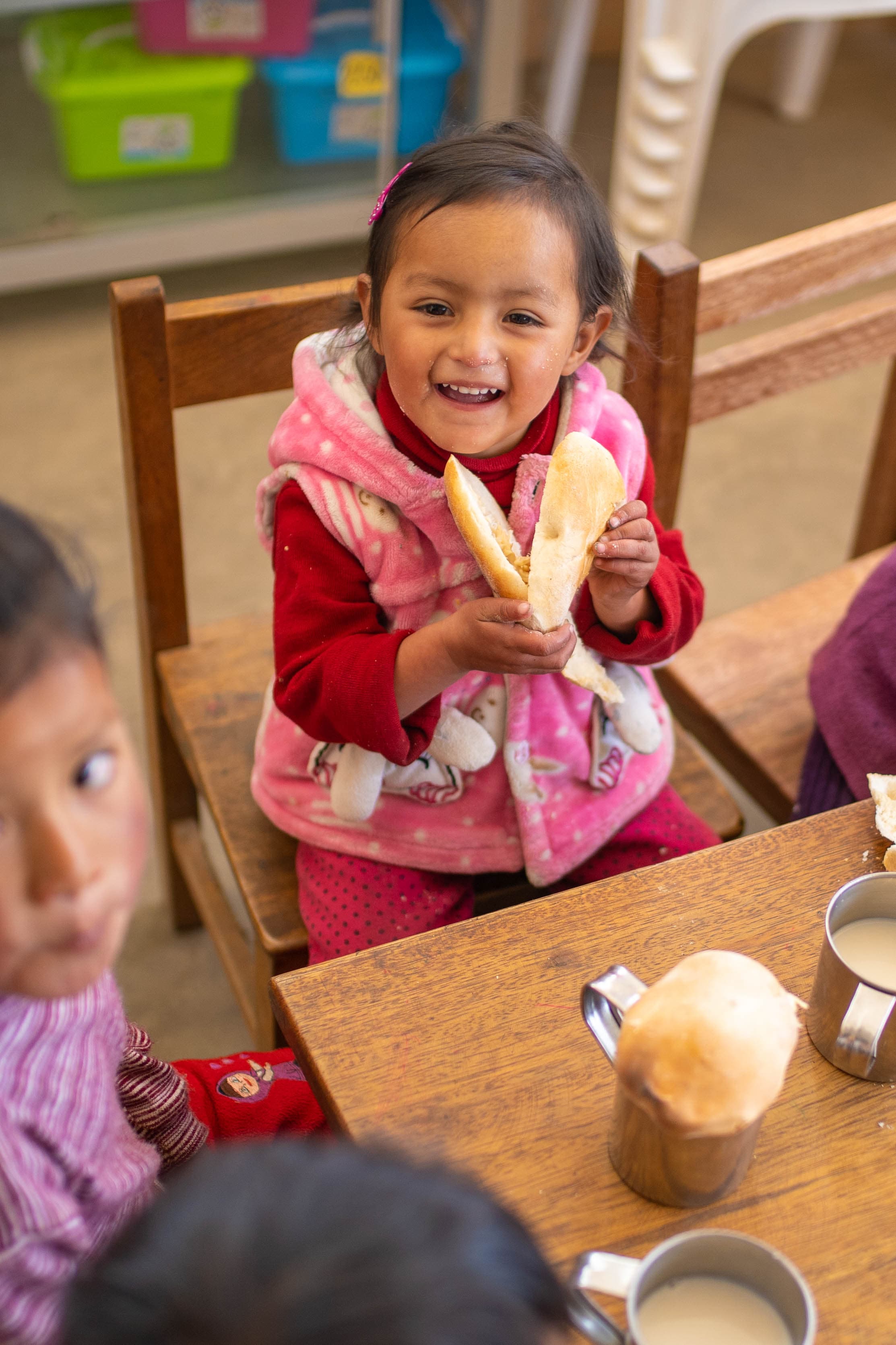 Little girl smiles as she holds a sandwich while sitting on a chair in her classroom.