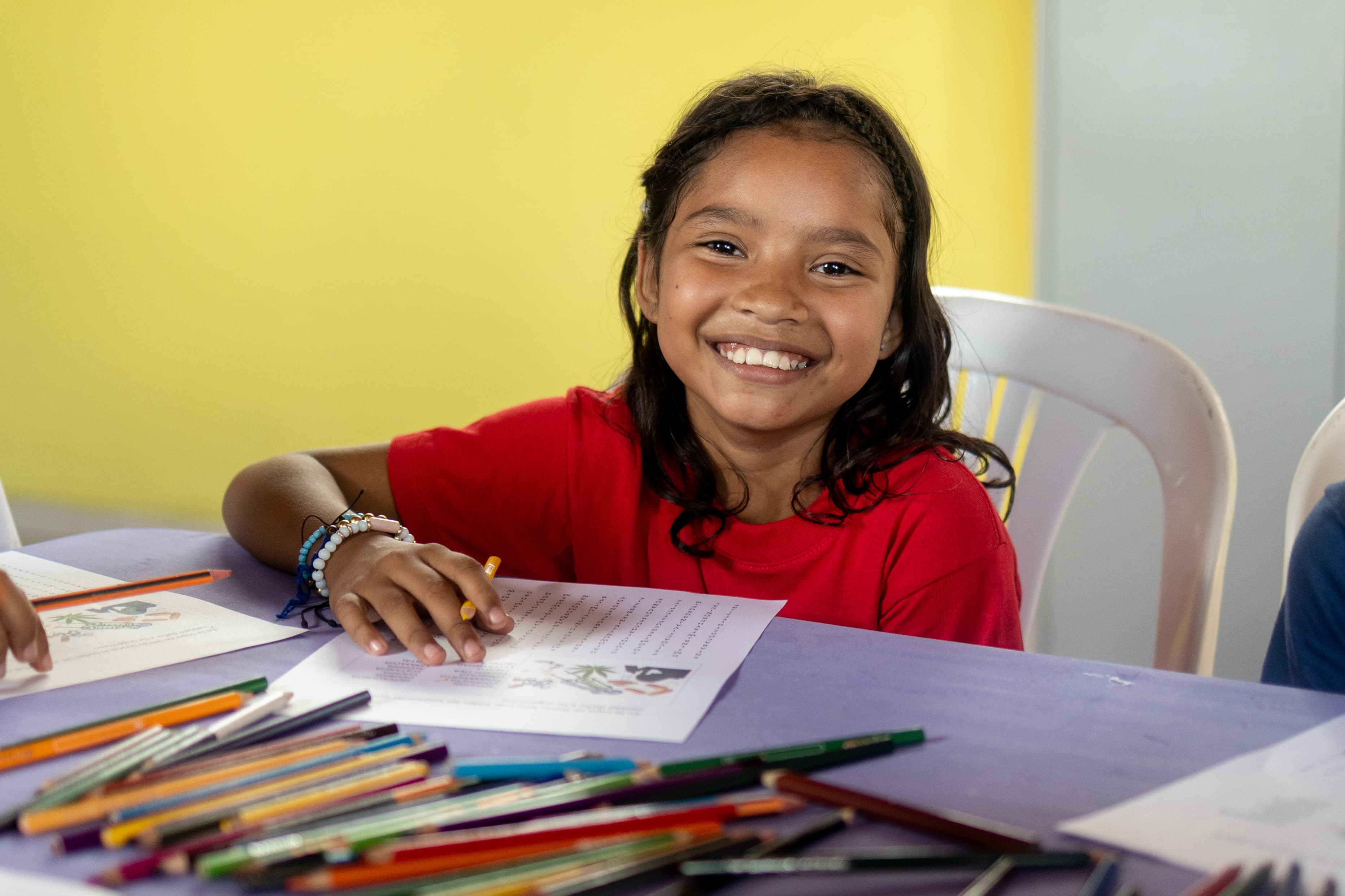 Girl in red shirt smiles, sits at table writing a letter.