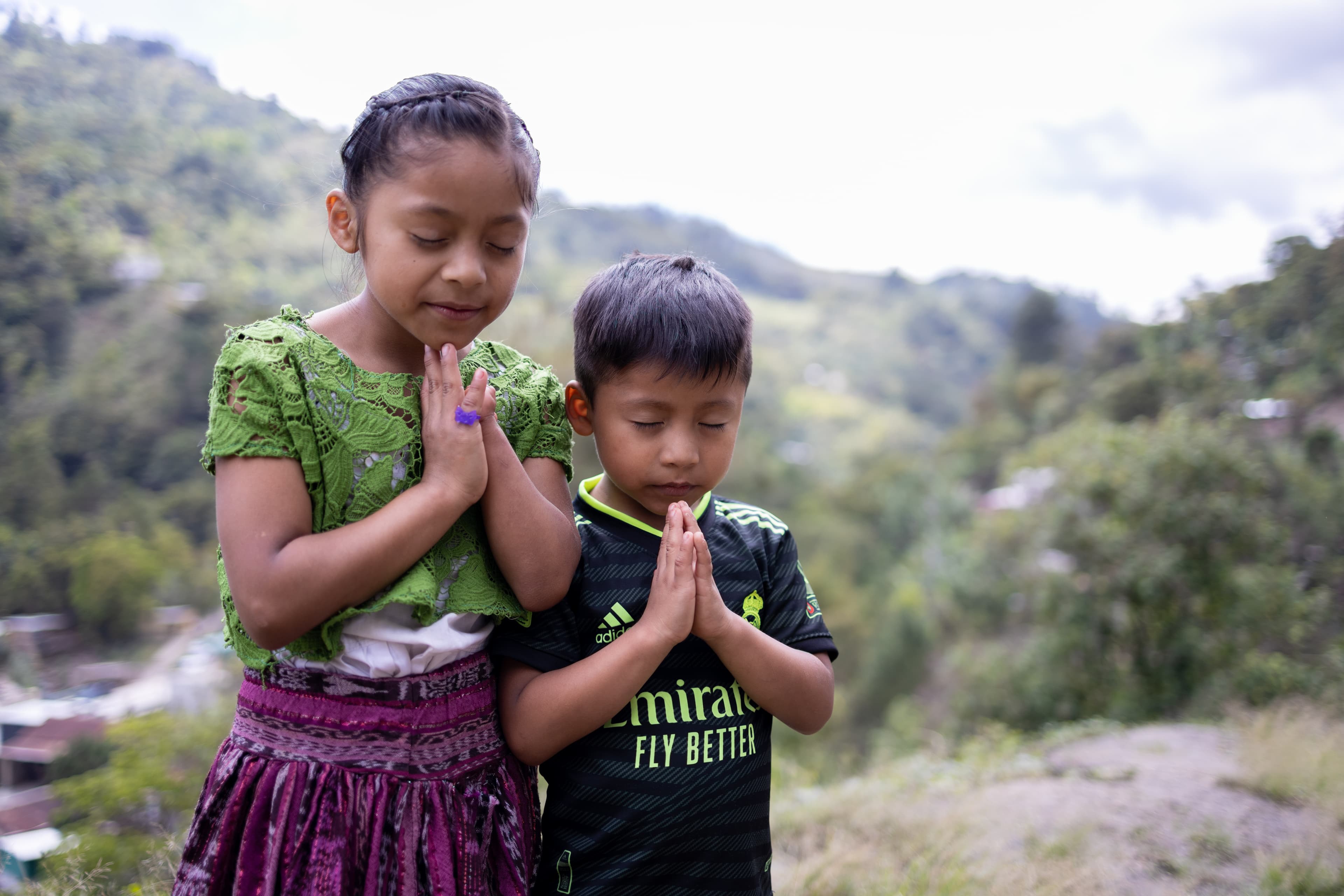 A sister and brother are standing together outdoors with their eyes closed and hands clasped in prayer.