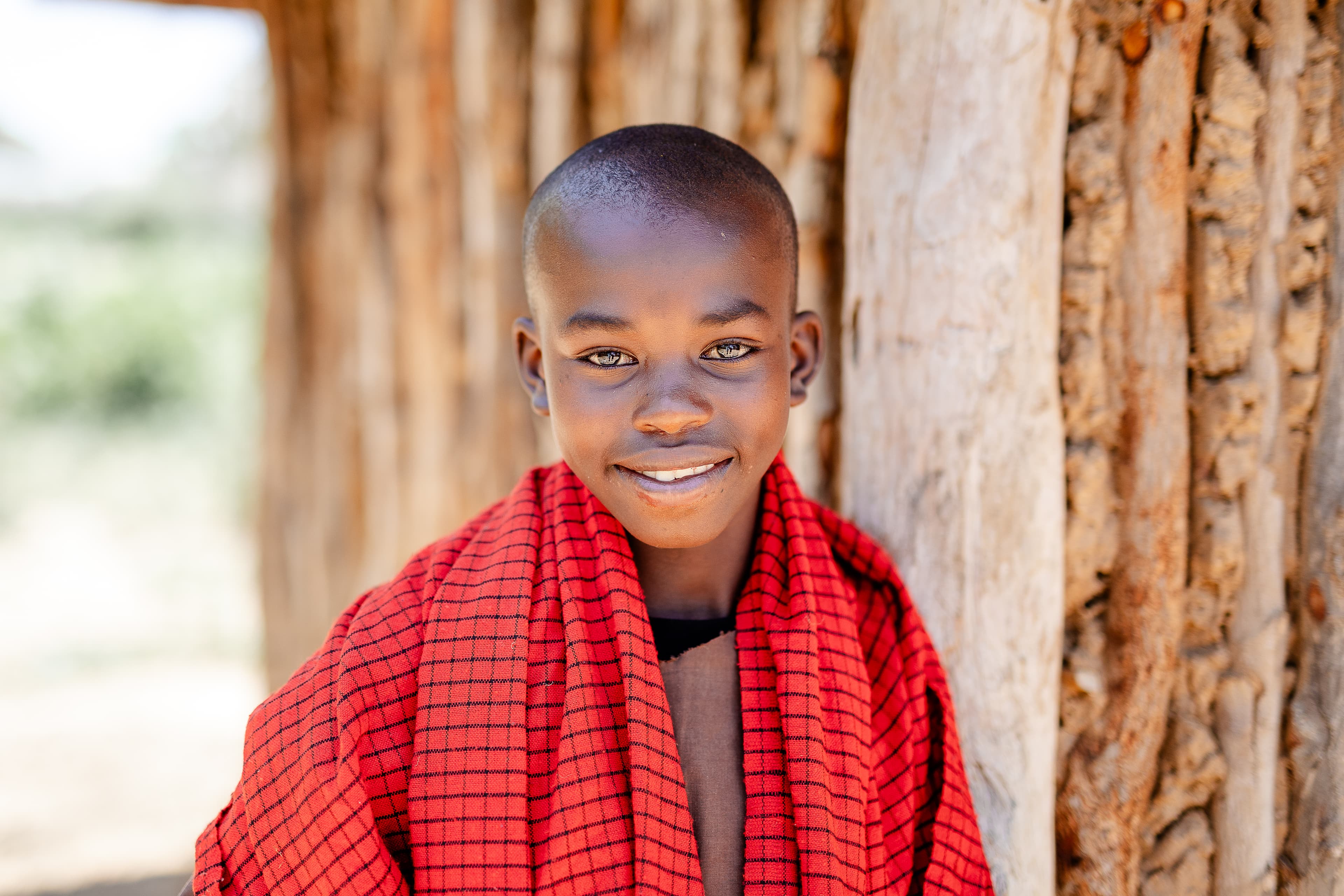 A boy dressed in a brown costume with red scarf stands in front of a wood and clay house, smiling.