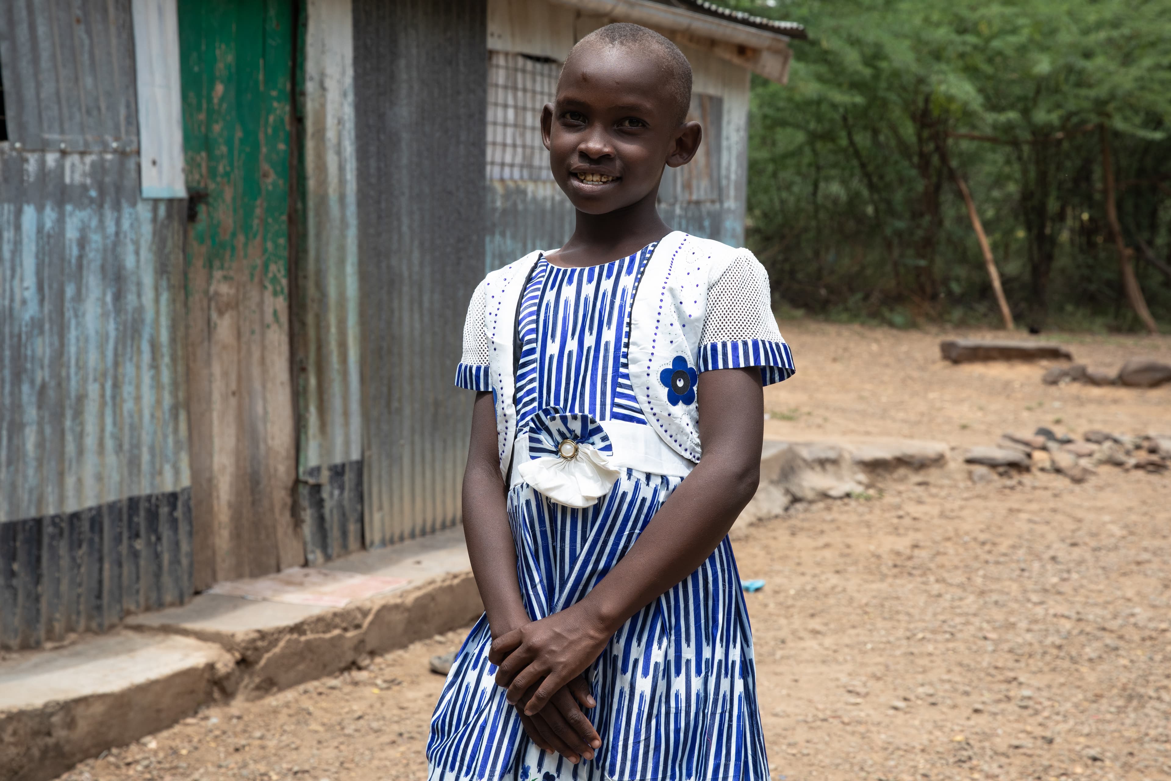 A young girl is standing with her hands clasped and smiling at the camera.