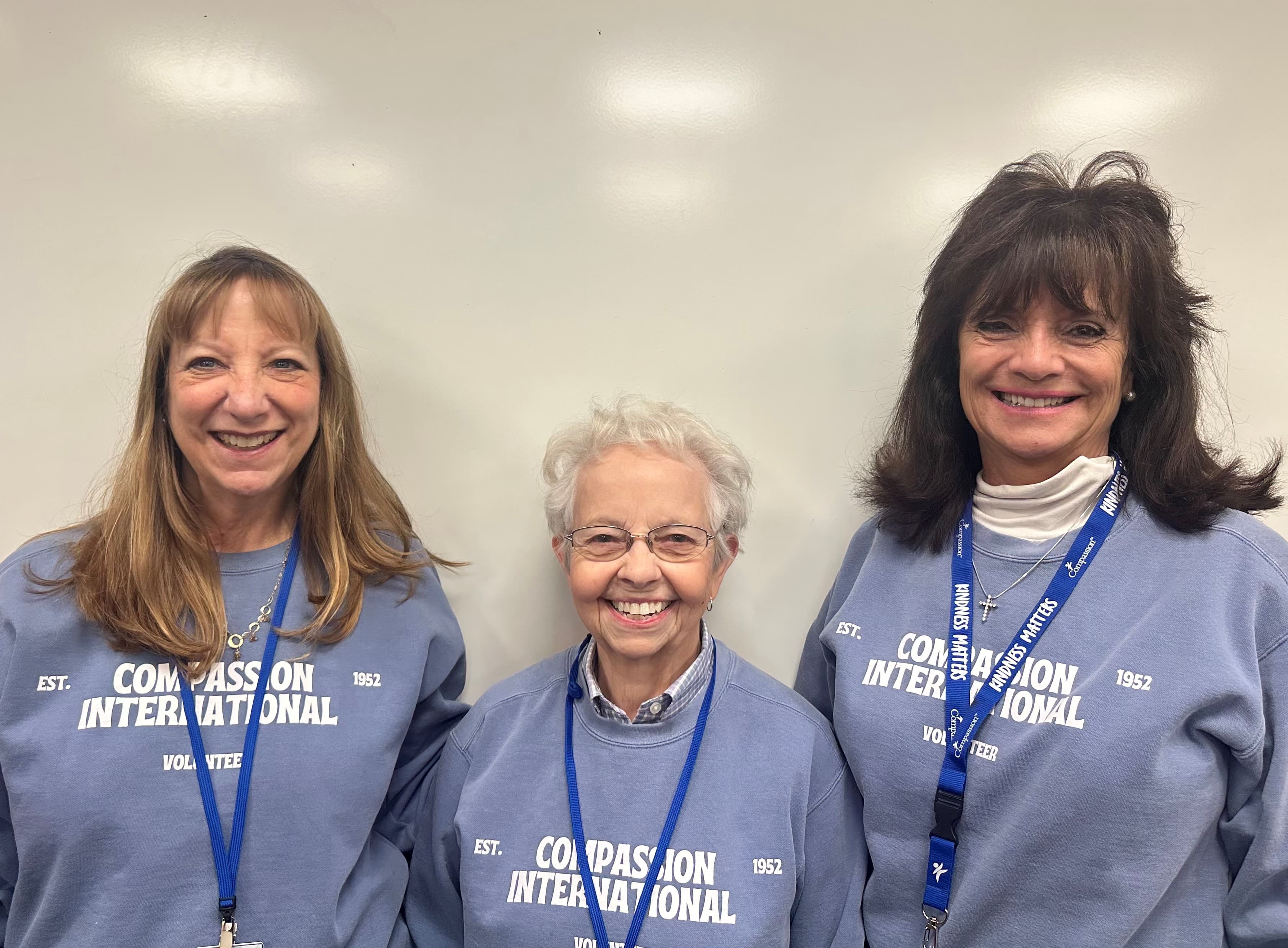 Three women are standing together wearing Compassion International sweatshirts.