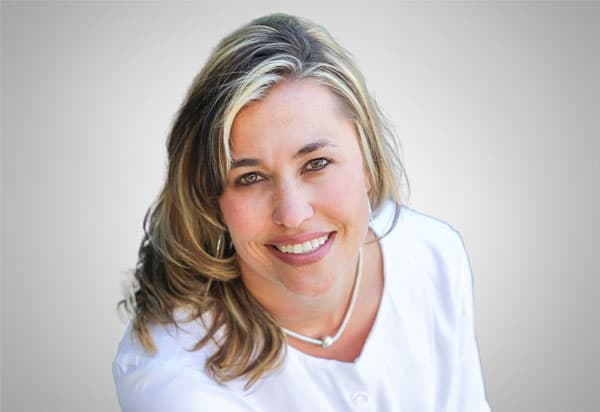 A woman in a white blouse and silver necklace is smiling, with a grey background with two black lines behind her.