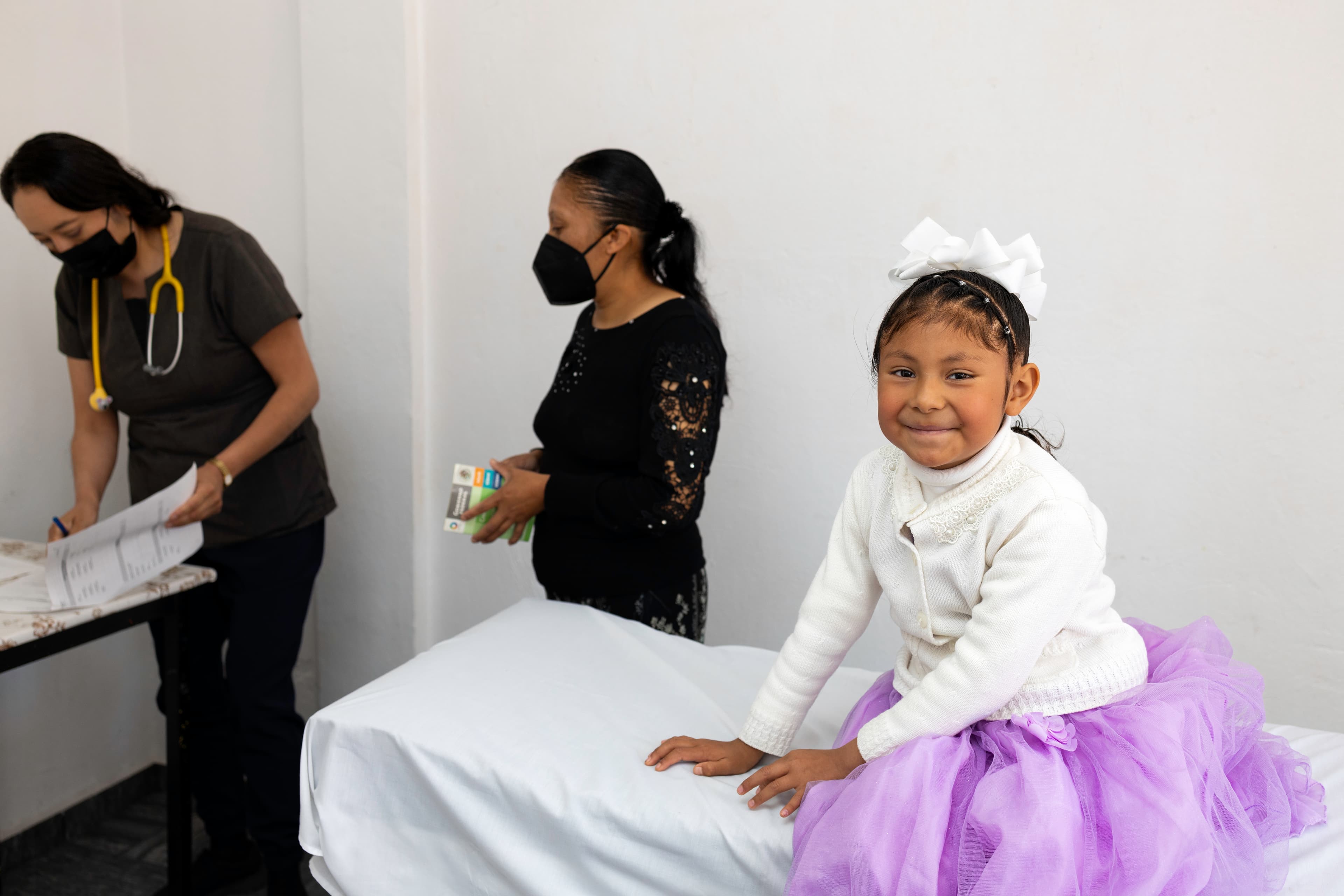 A young girl wearing a purple dress sits on a hospital bed while smiling.