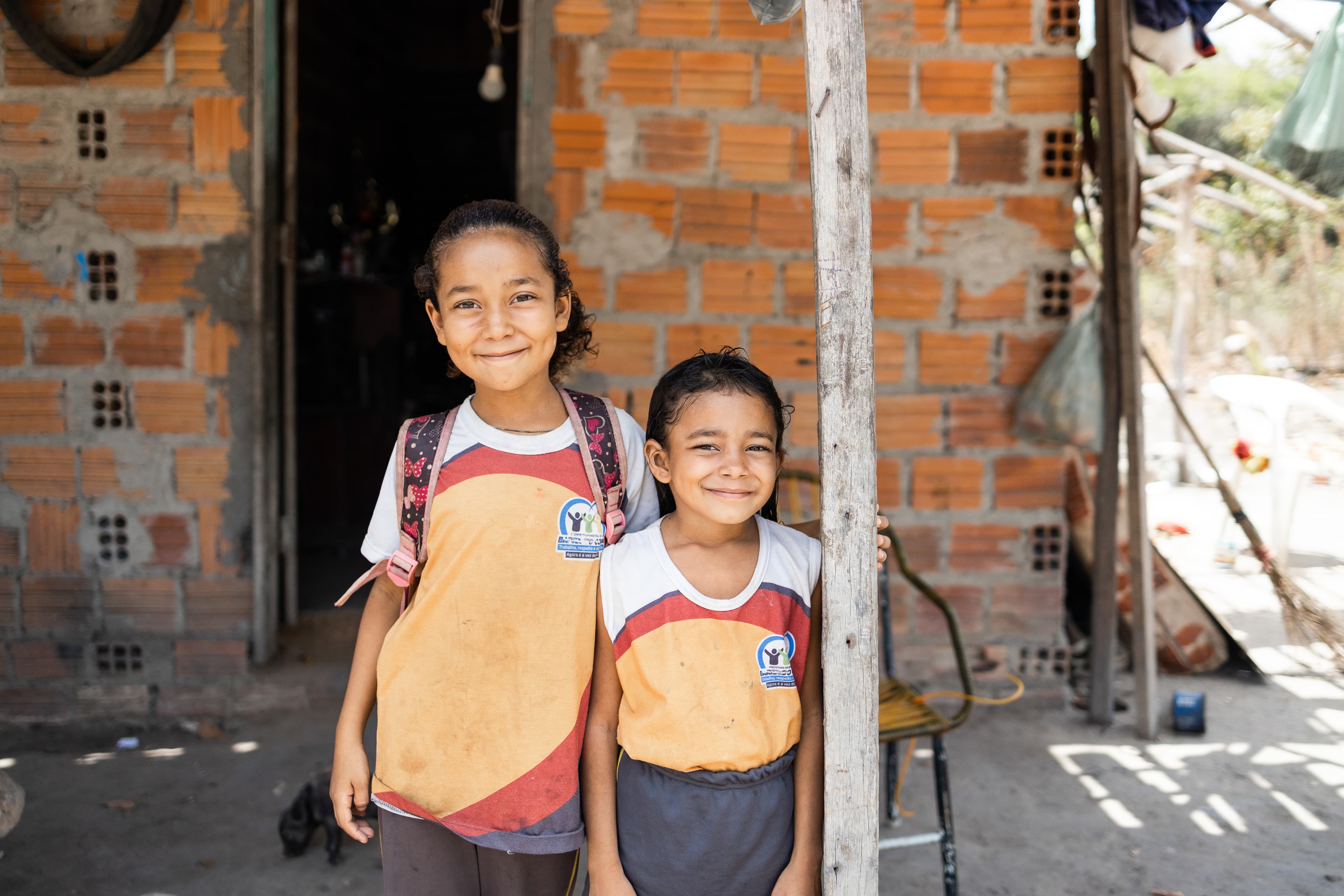 Two young girls stand outside of their home smiling and wearing their school uniforms.