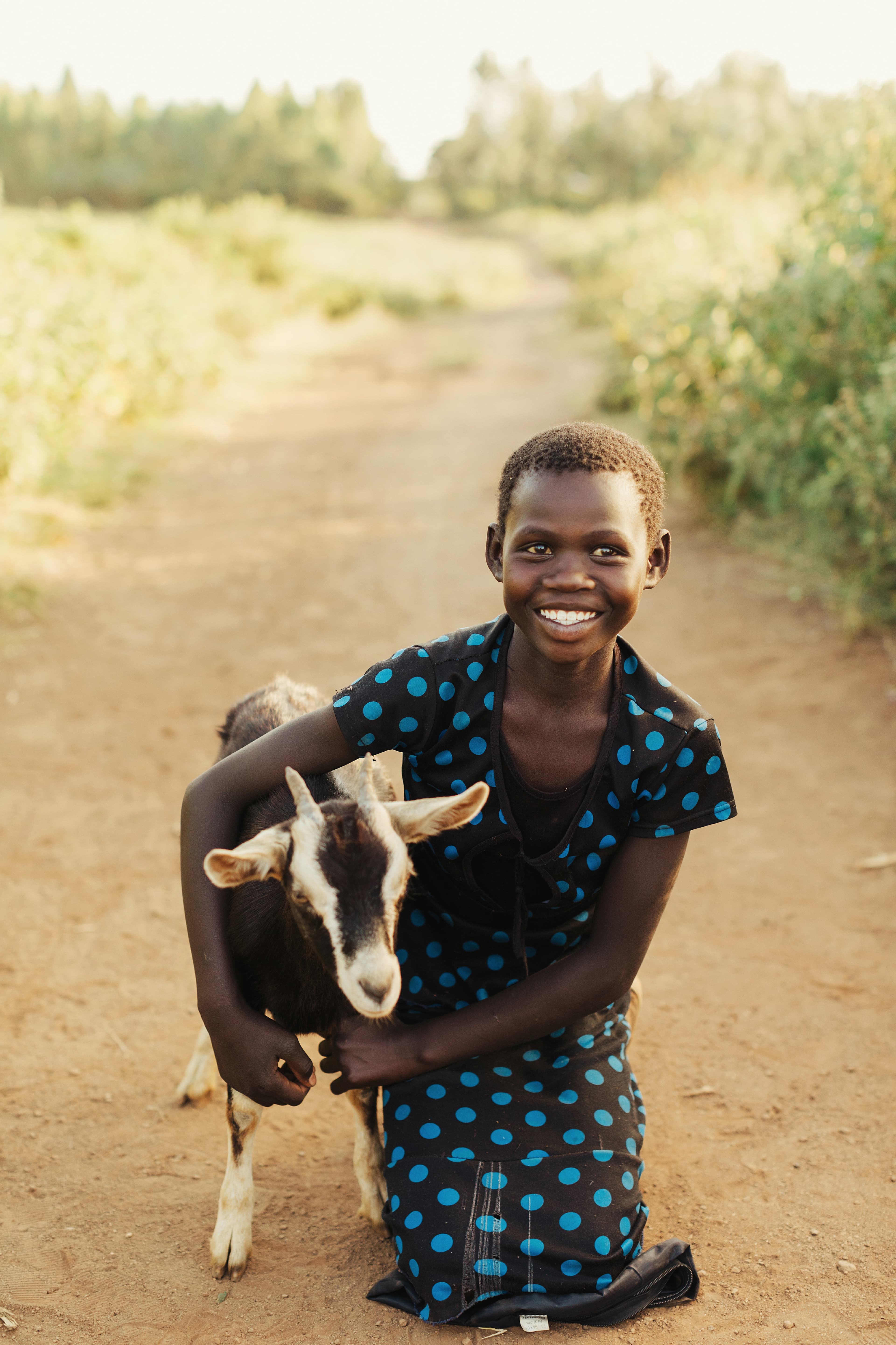 A young girl is kneeling on the ground holding a goat and smiling.