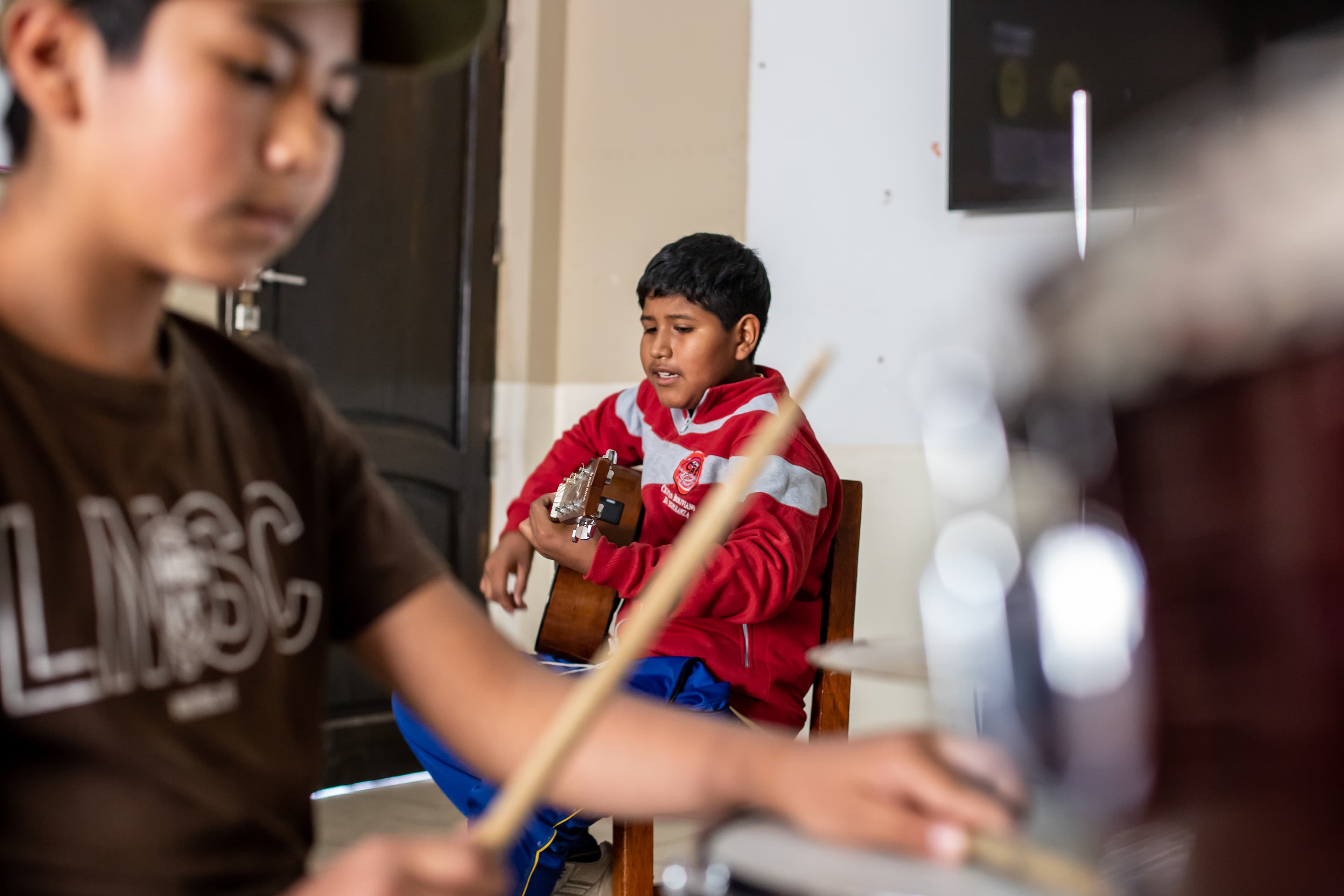 An older boy in a red sports jacket sings while playing guitar. Another boy plays drums in the foreground.