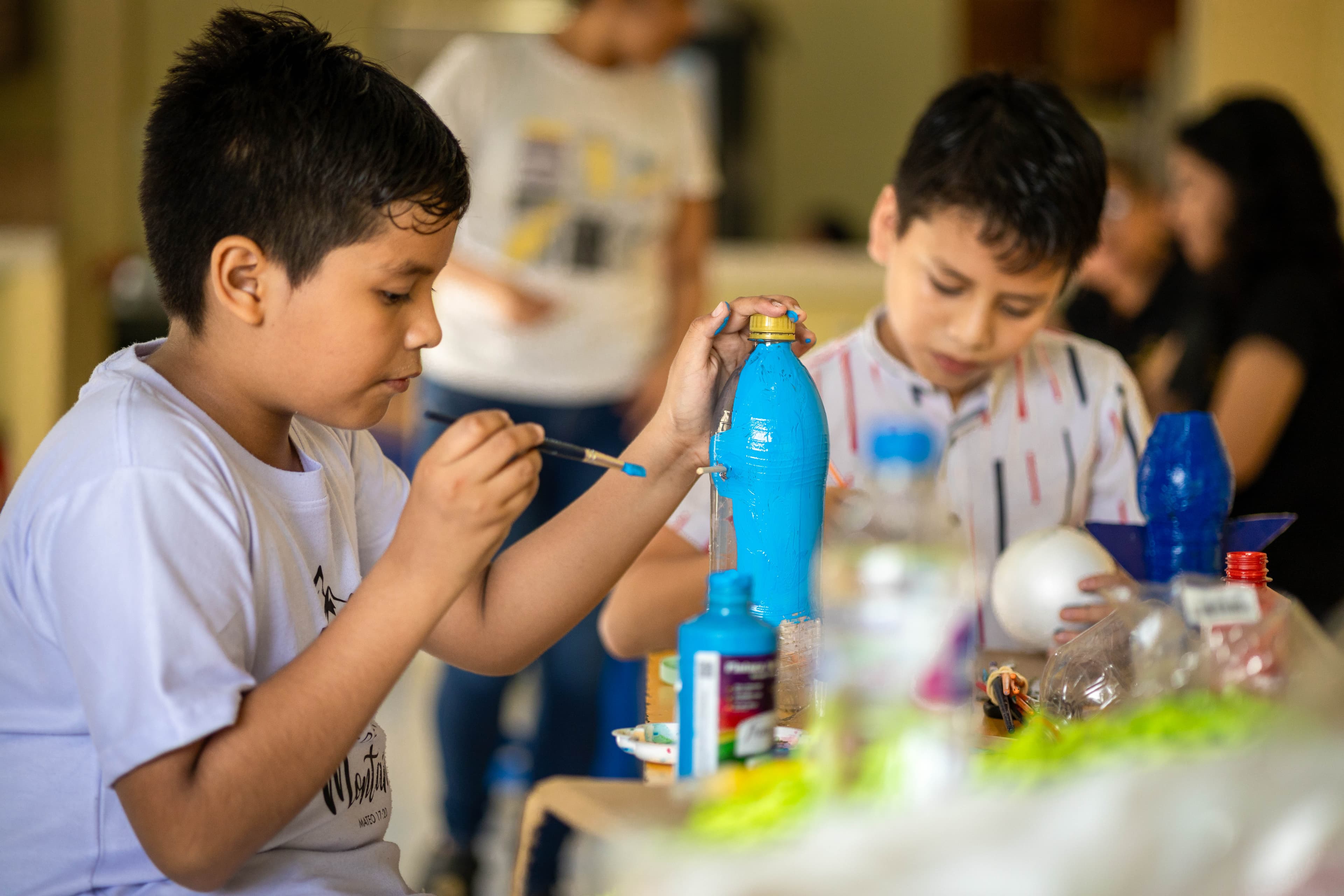 12 year old Jinsop holds a paintbrush and paints a water bottle bright blue. there are recycled materials and art supplies on the table around him. behind him is 9 year old Santi, who is concentrating on paiting his project.