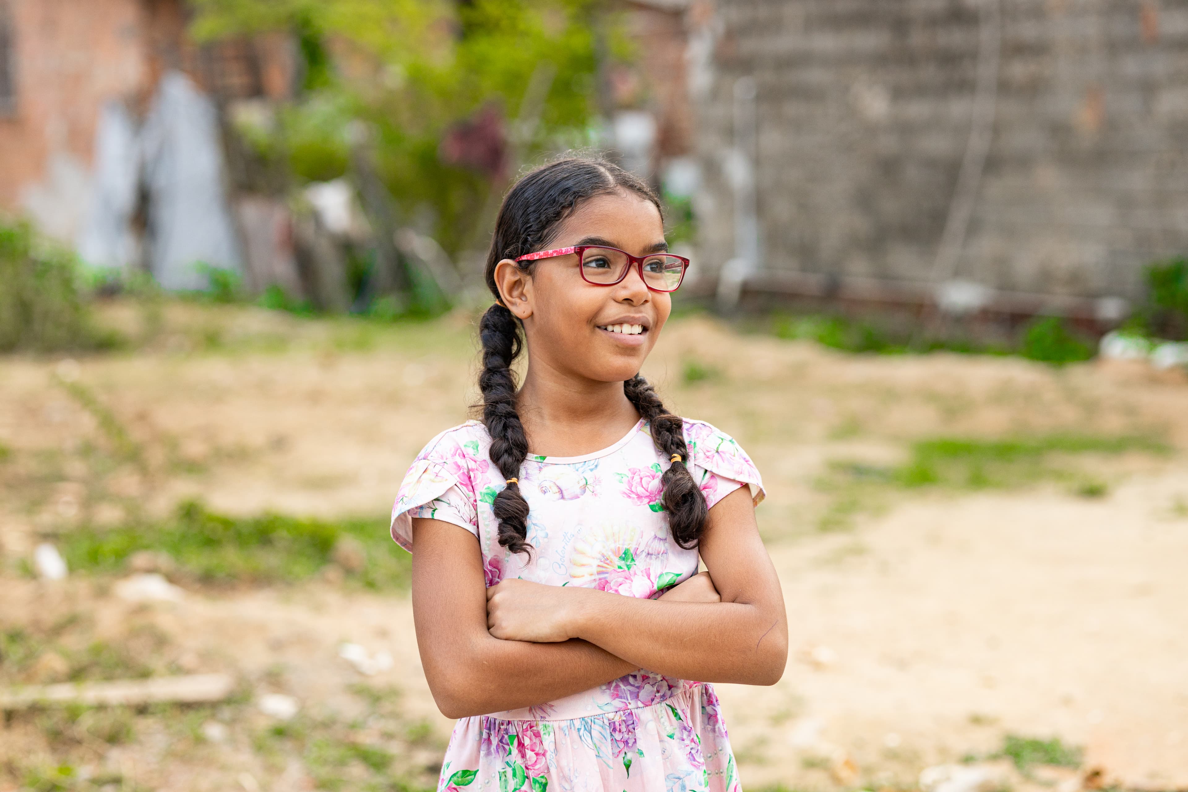A young girl wearing a floral dress and glasses crosses her arms and smiles.