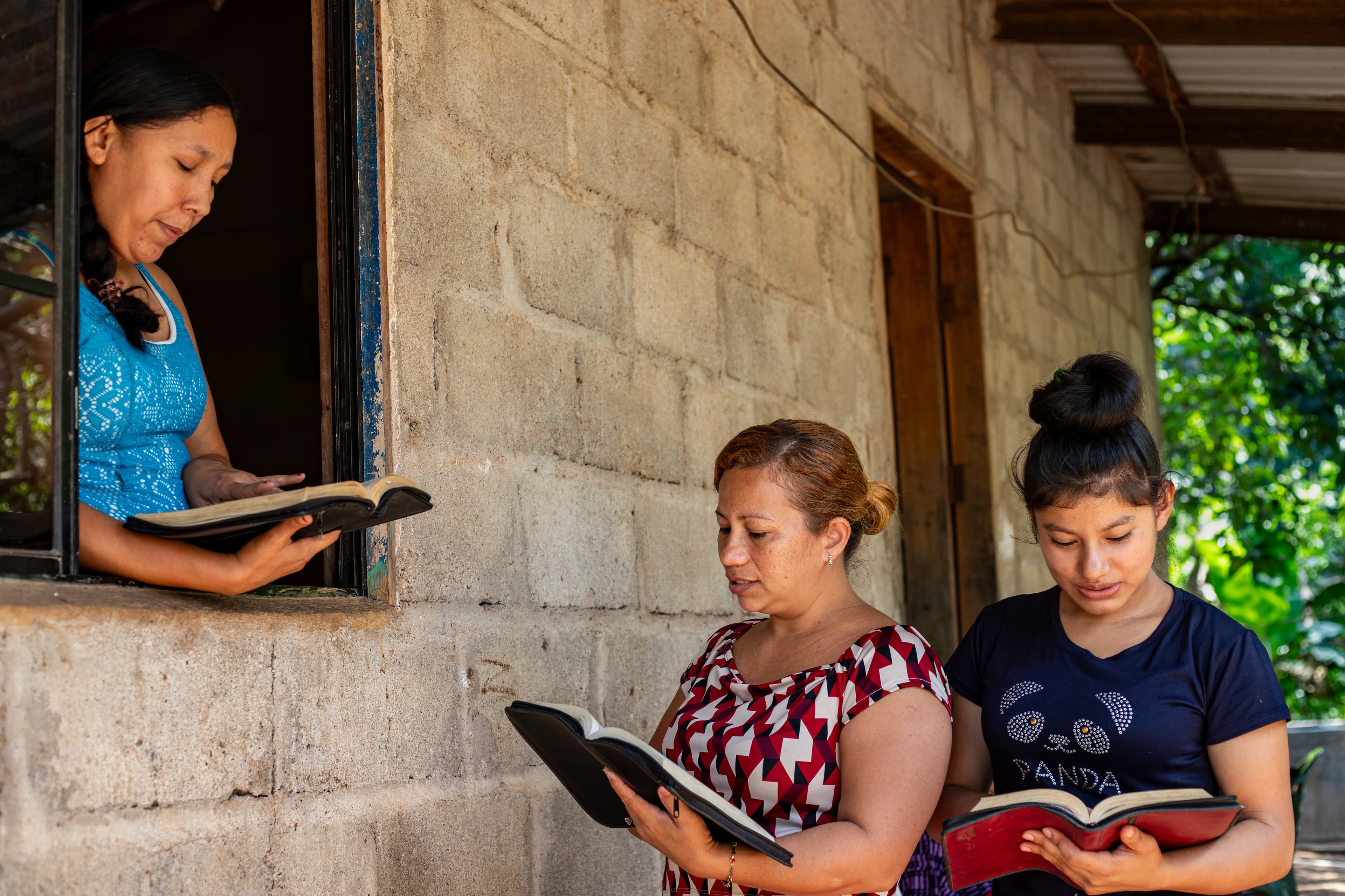 Three young women read their Bibles together outside of a block building.