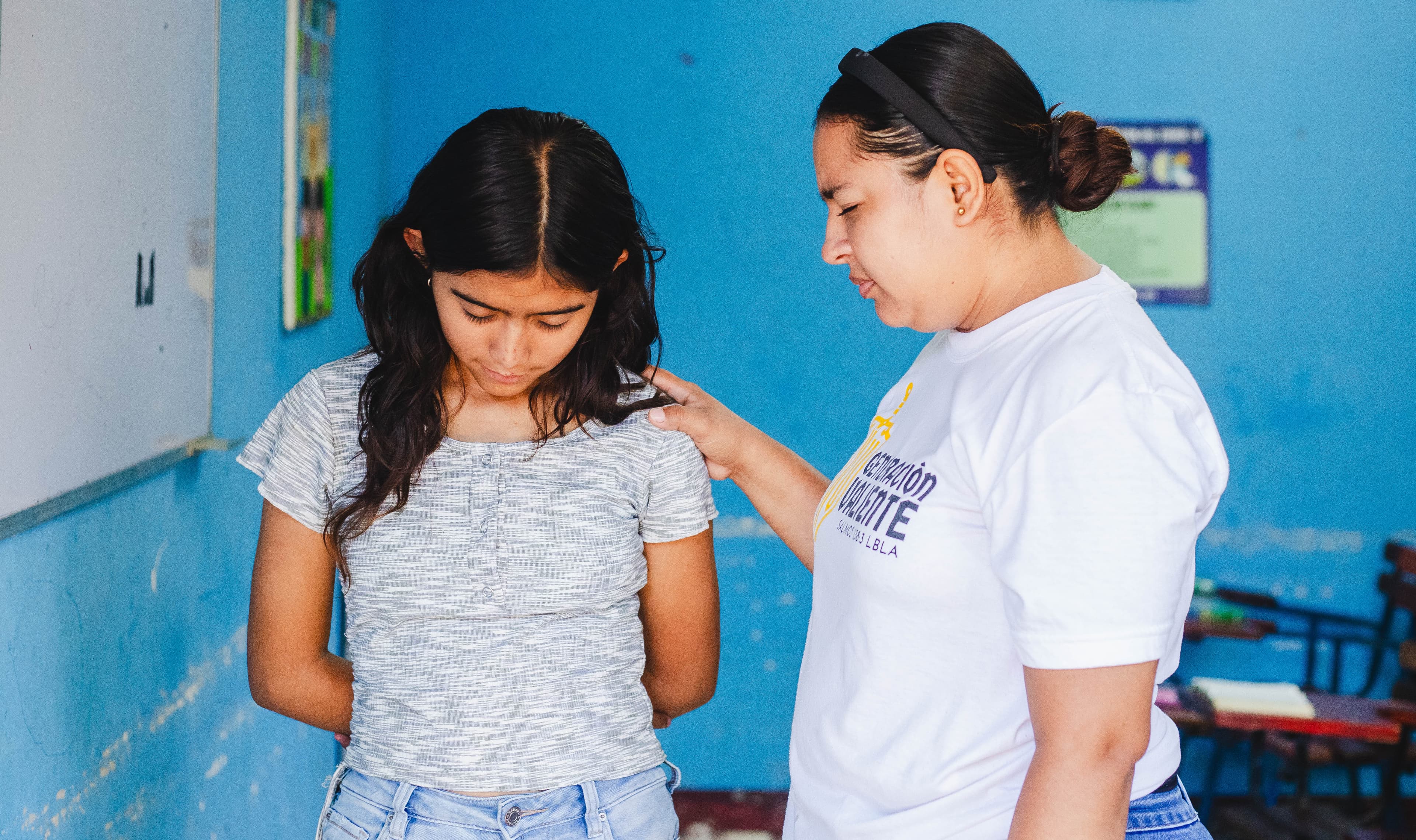 A young woman places her hand on the shoulder of a teen girl as they pray.