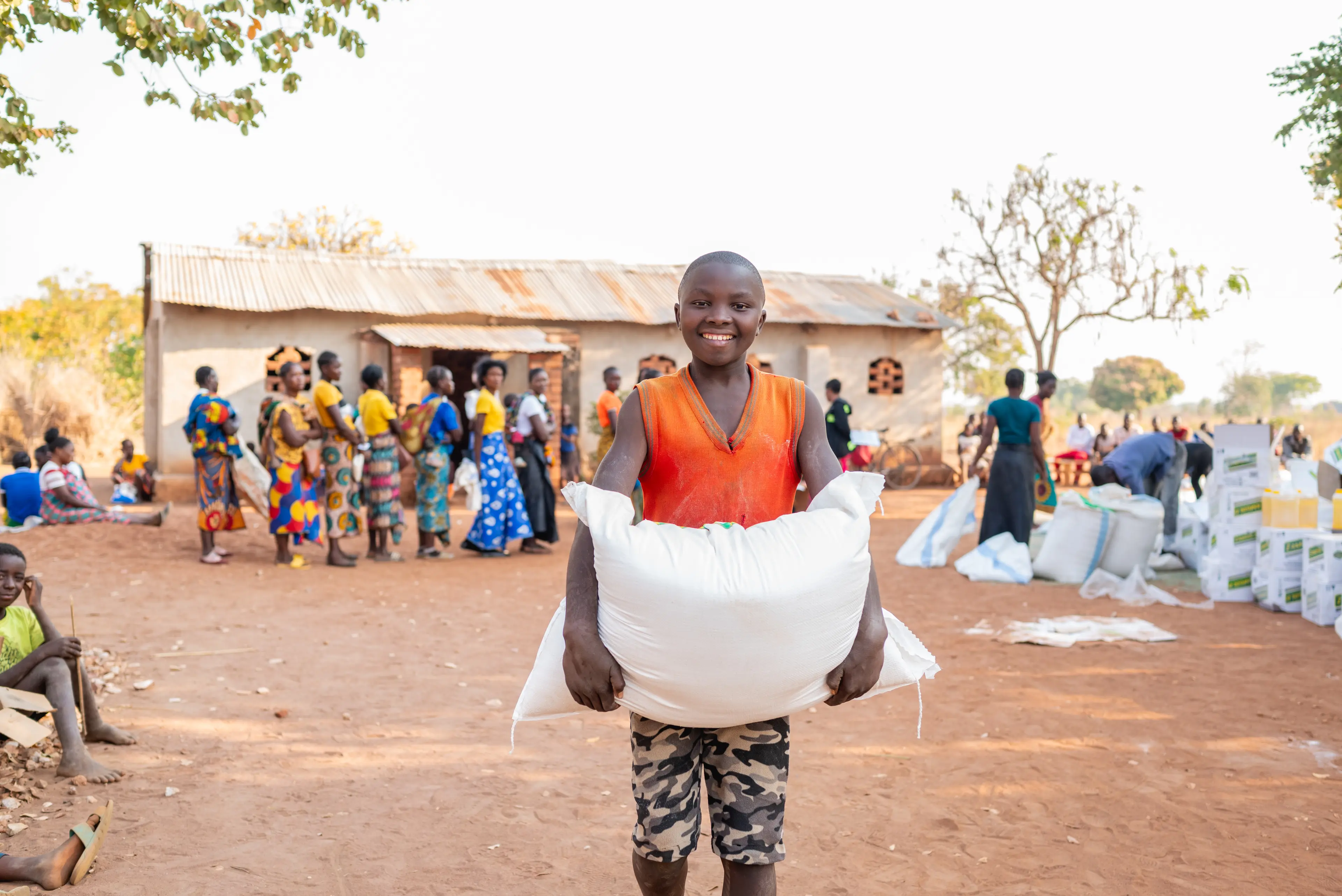 A young African boy wearing a bright orange shirt holds a large bag of food while smiling.