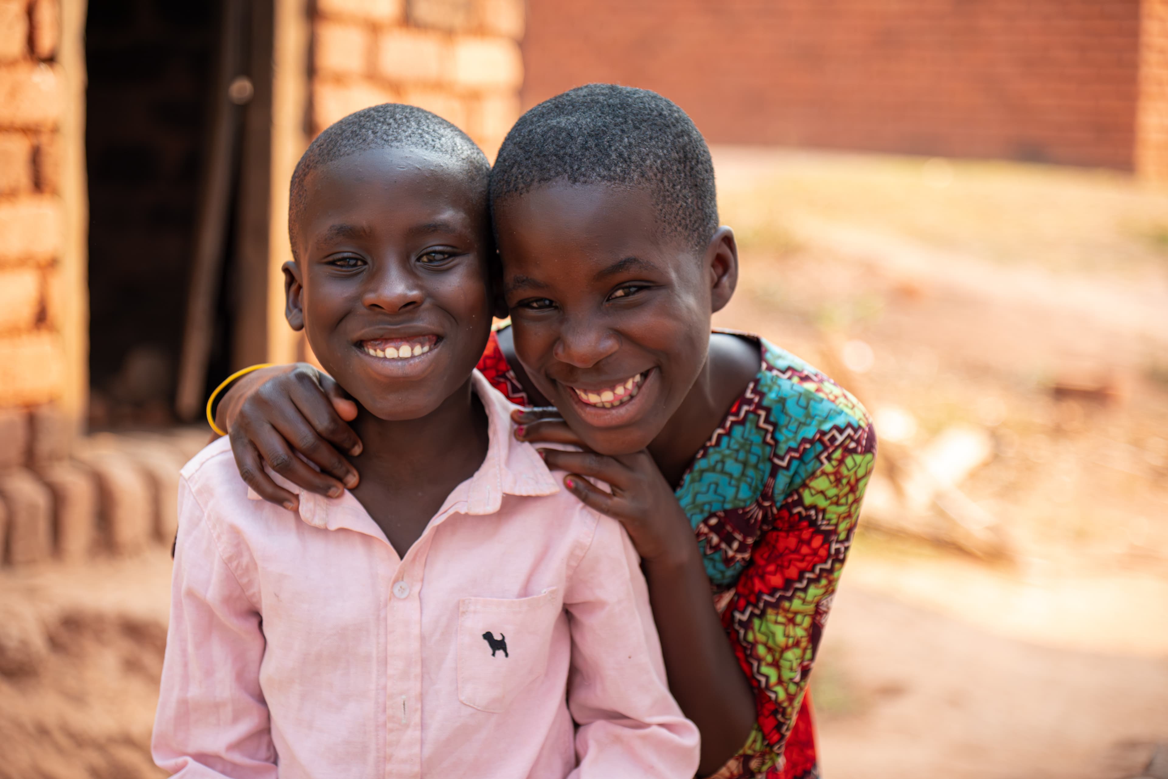 A boy in a pink oxford shirt sits with his sister just behind him and they both smile at the camera, and in the background are brick buildings.